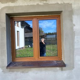 WINFOX GROUP - Brązowe okno dwuskrzydłowe w surowym murze, z widoczną pianką montażową. W szybie odbija się budynek i osoba robiąca zdjęcie.