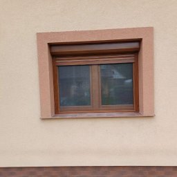 WINFOX GROUP - Brązowe okno z roletą zewnętrzną w beżowej elewacji domu. Widoczny fragment ceglanej podmurówki i lampa solarna. Ujęcie z bliska, światło dzienne.