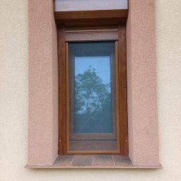 WINFOX GROUP - Drewniane okno z moskitierą i roletą zewnętrzną w brązowej ramie, osadzone w elewacji o ciepłej kolorystyce. Widok z zewnątrz, odbicie drzewa w szybie.