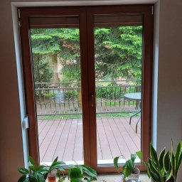 WINFOX GROUP - Drewniane dwuskrzydłowe okno balkonowe z widokiem na taras i zieleń. Wnętrze zdobią rośliny doniczkowe. Widoczne rolety zwinięte u góry okna.