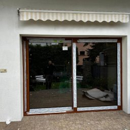 WINFOX GROUP - Brązowe, przesuwne drzwi tarasowe z widocznym odbiciem osoby, zamontowane w białej ścianie z markizą. Na zewnątrz kostka brukowa i zieleń.