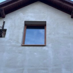 WINFOX GROUP - Drewniane okno z widokiem na niebo, osadzone w surowej, szarej elewacji domu z brązowym dachem. Widoczne ślady po pracach budowlanych.