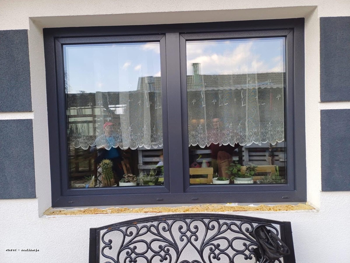 Szare okno PCV z firanką w domu o białej elewacji z szarymi wstawkami. Na parapecie wewnętrznym rośliny w doniczkach. Na zewnątrz ozdobny element metalowy.