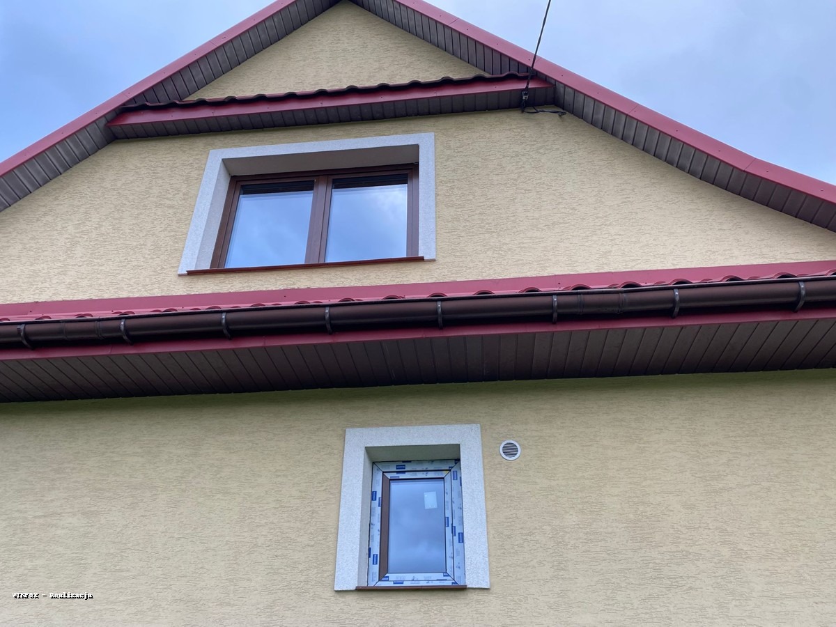 Elewacja domu z dwoma oknami o różnej wielkości i kolorze ram, brązowe rynny i czerwone wykończenie dachu. Okno na poddaszu z brązową ramą.
