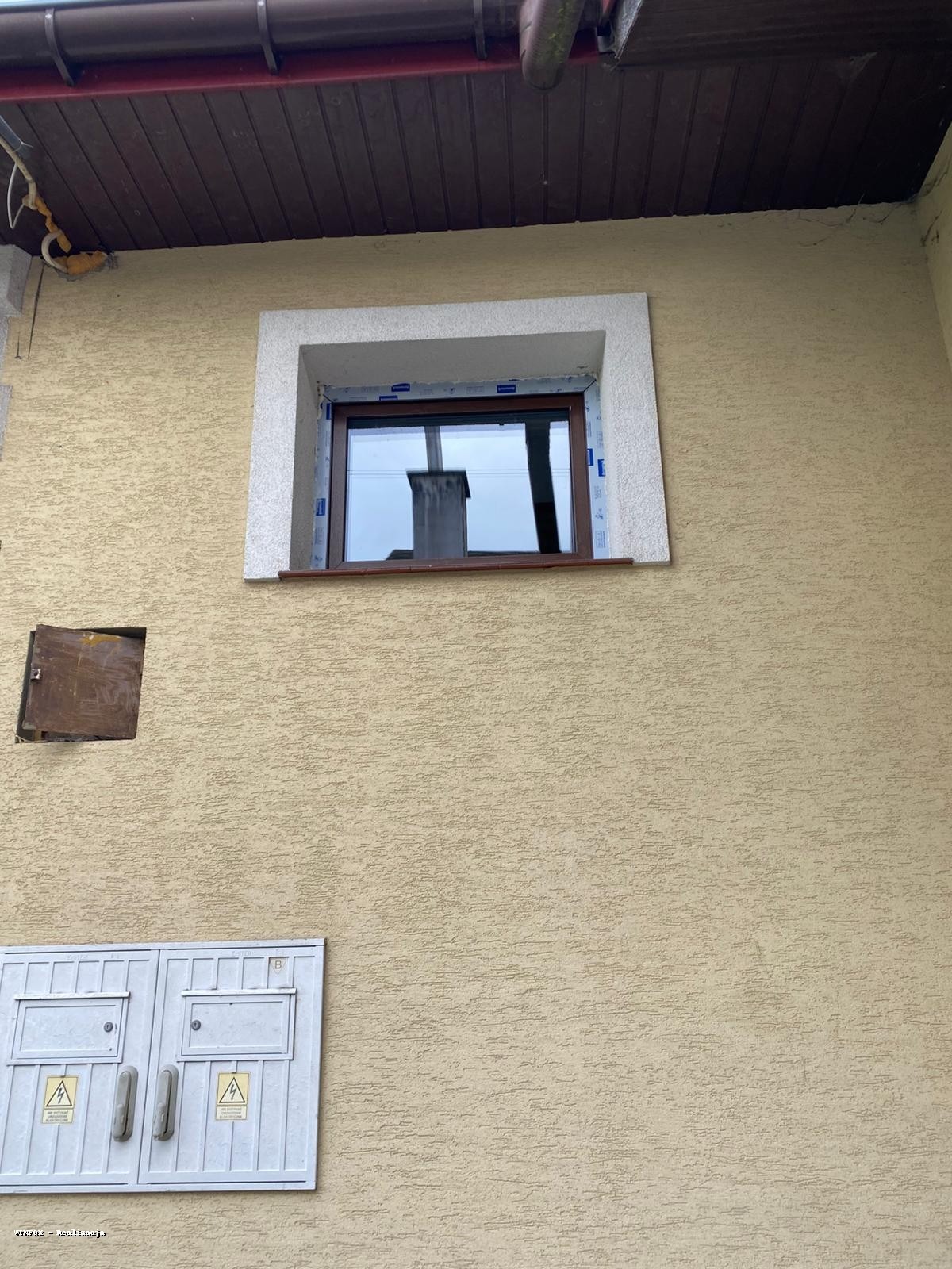 Brązowe okno z białą ramą osadzone w elewacji budynku o beżowej strukturze baranka. Widoczna skrzynka elektryczna i fragment rynny.