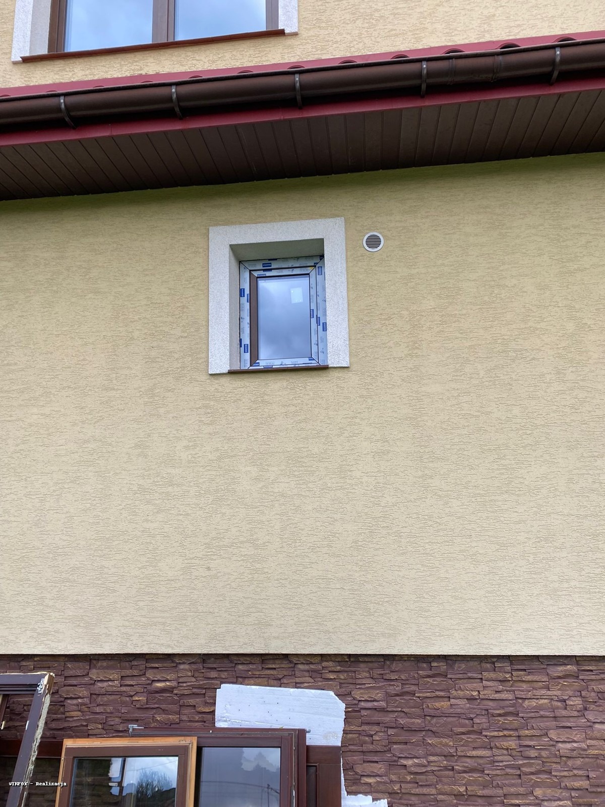 Elewacja budynku z zamontowanym oknem w białej ramie. Na dole leżą stare ramy okienne. Widoczny fragment dachu z rynną. Tynk w kolorze beżowym.