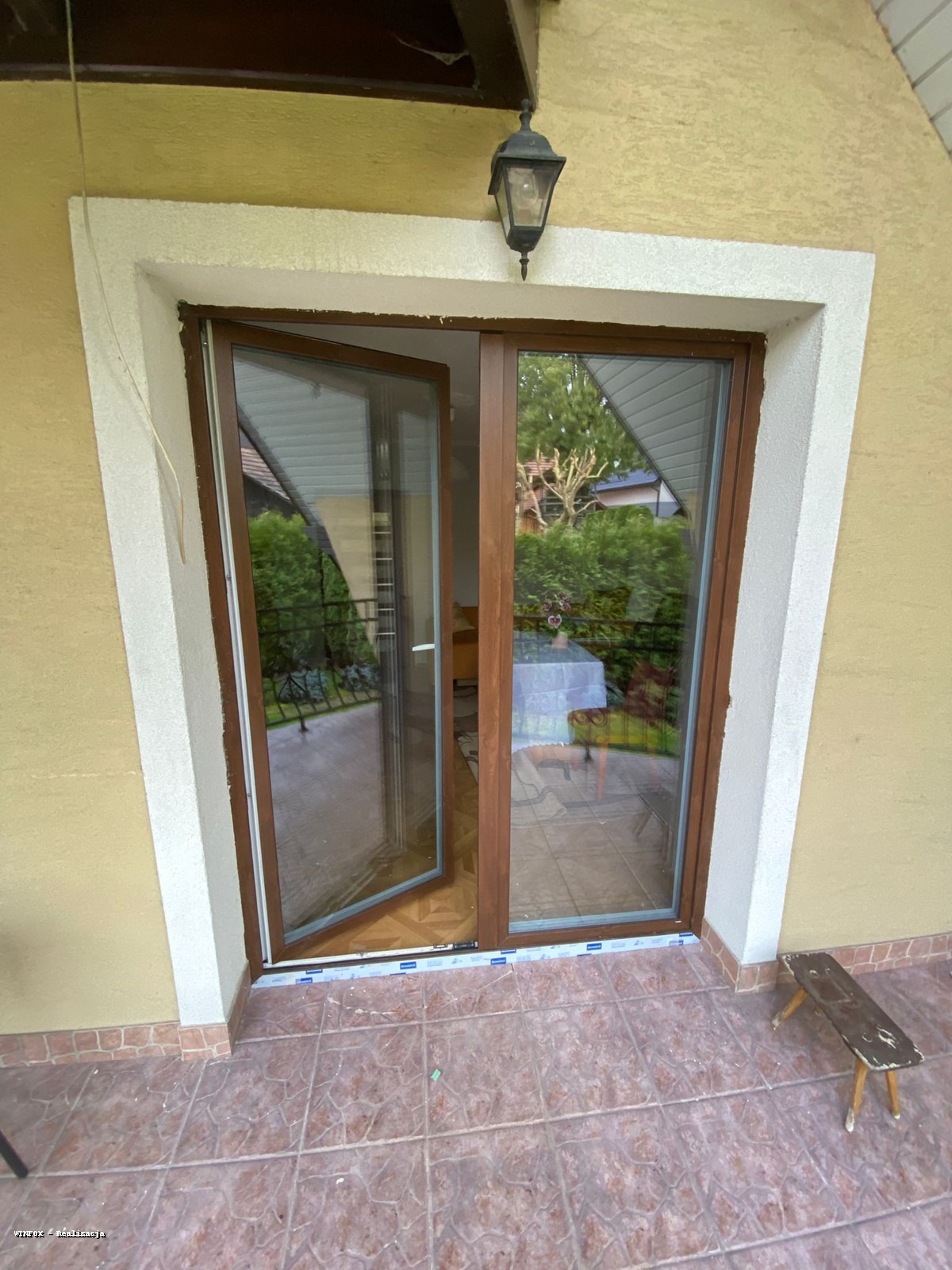 Rozwarte brązowe drzwi balkonowe w Działoszycach, widok na zieleń ogrodu. Stolarka z profili PCV, białe obramowanie, lampa zewnętrzna nad wejściem.