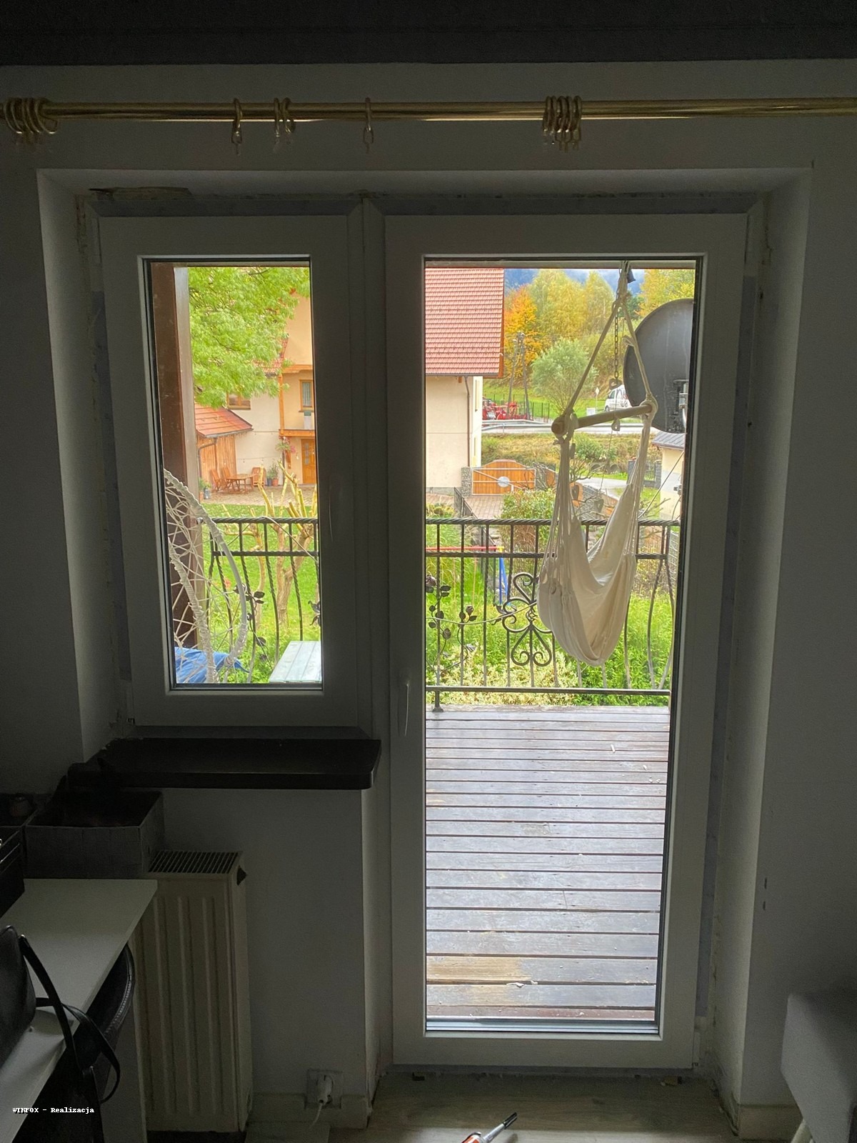 Białe okno balkonowe z widokiem na taras i ogród. Na balkonie wiszący hamak. Widoczne detale wykończenia wnętrza. Ujęcie z wnętrza pomieszczenia.