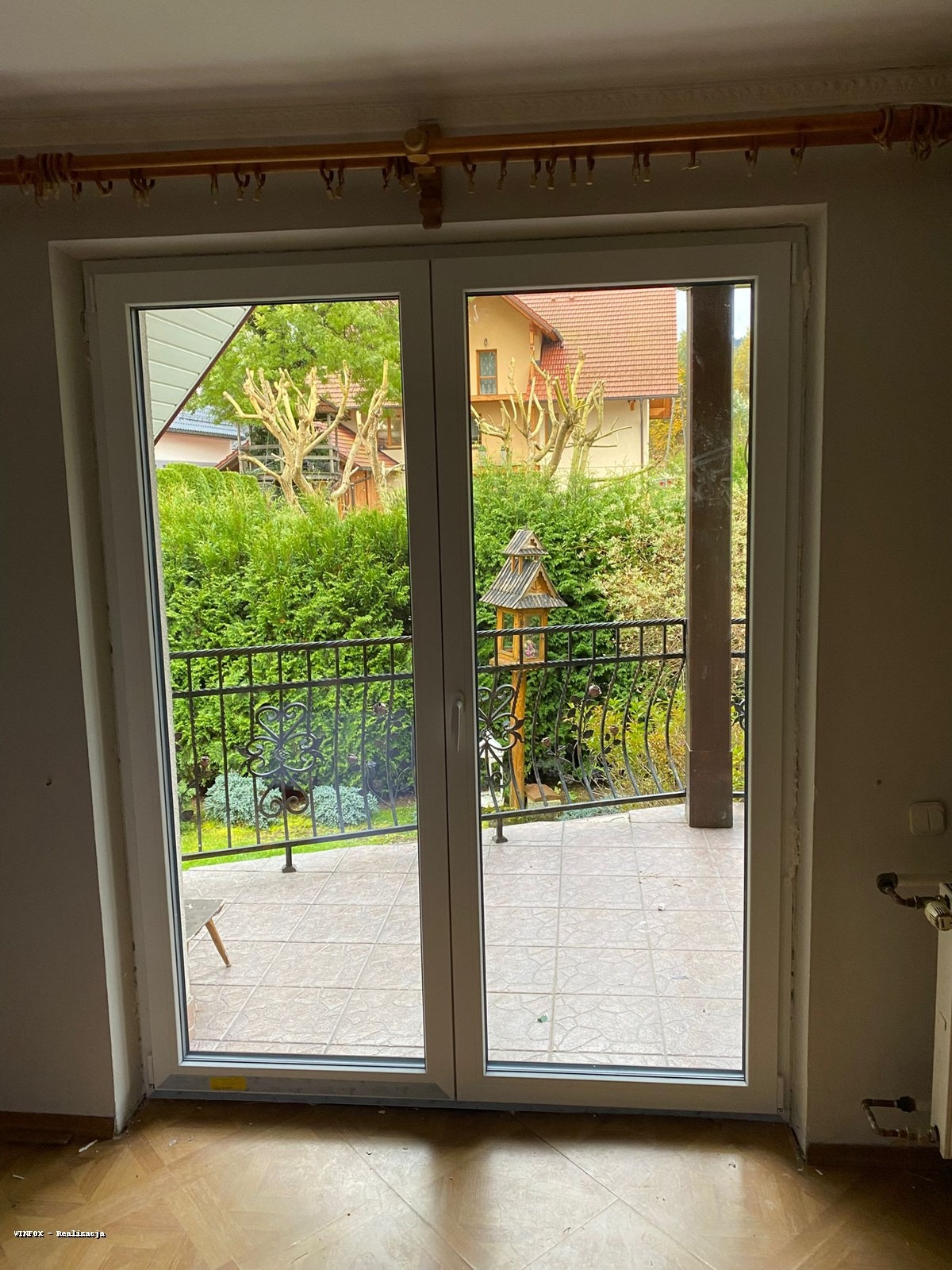 Białe, dwuskrzydłowe drzwi balkonowe PCV z widokiem na balkon z kutą balustradą, zielony ogród i domek dla ptaków. Widoczne drewniane wykończenia sufitu.