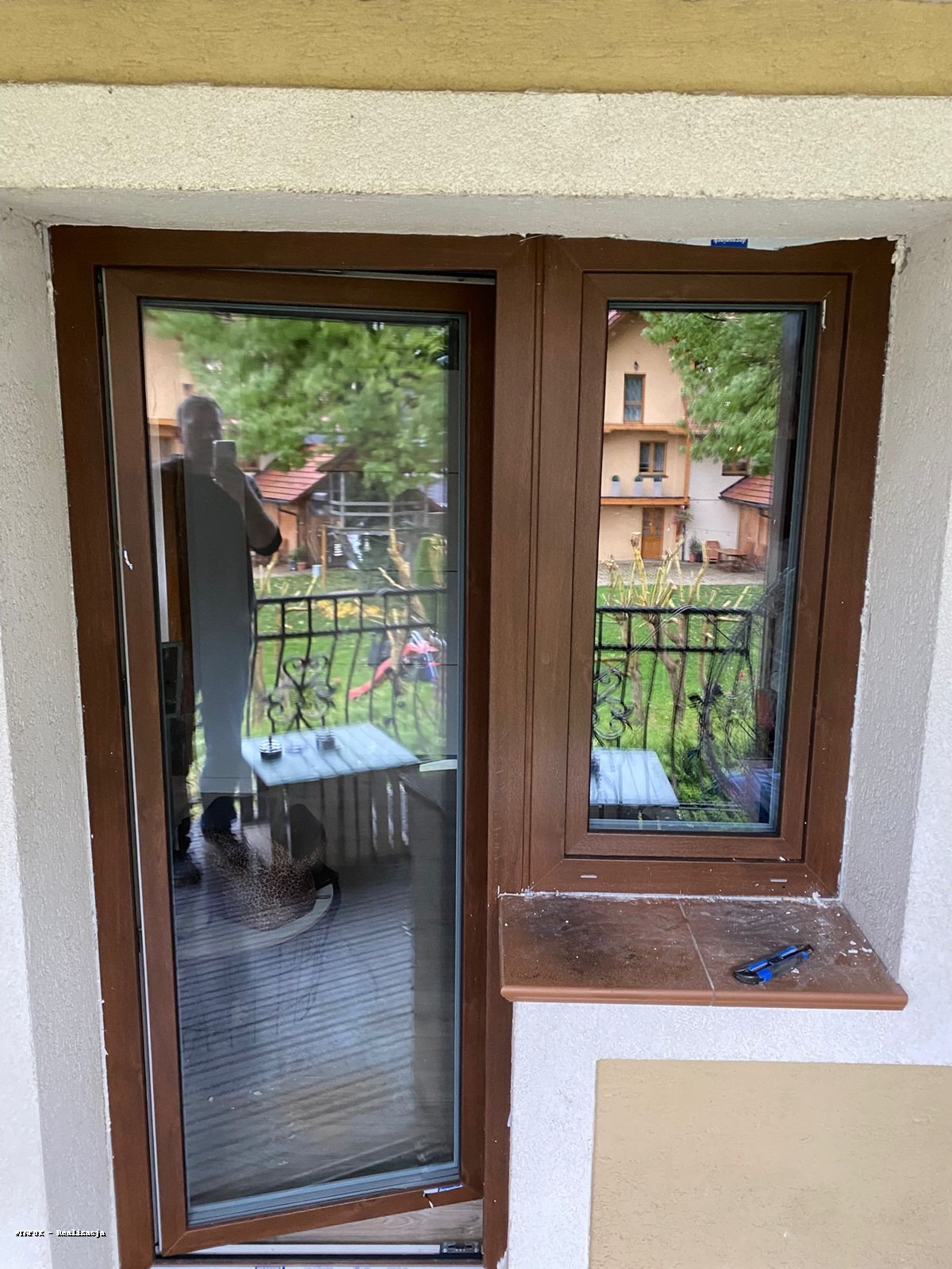 Brązowe okno balkonowe z uchylnym skrzydłem i małe okno obok, widok na balkon i ogród. Na parapecie leży nóż monterski. W szybie odbija się osoba.