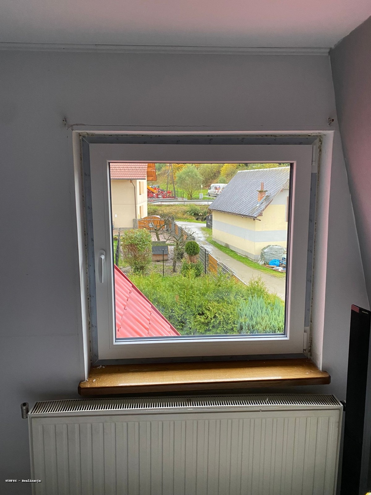 Białe okno z widokiem na ogród i domy w tle. Widoczny parapet i grzejnik pod oknem. Ujęcie z wnętrza pomieszczenia, okno w ramie z uszczelkami.