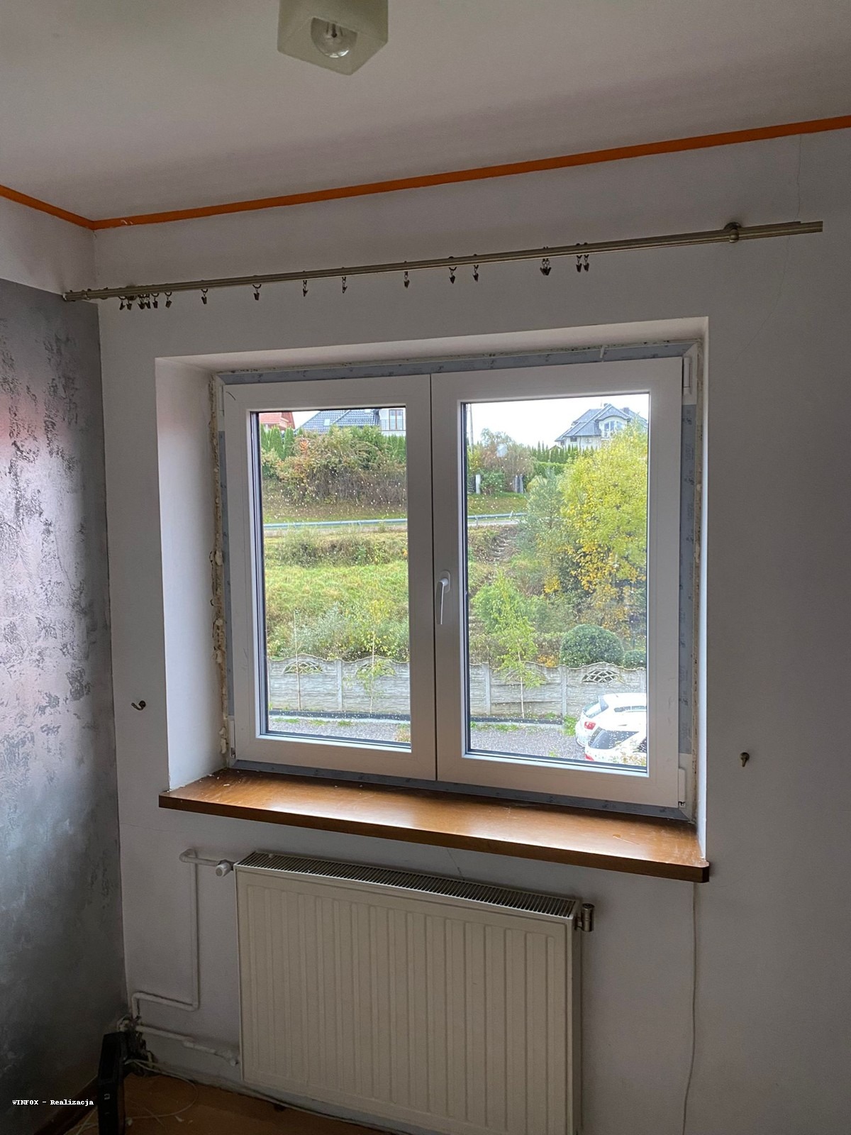 Montaż okna PCV z widokiem na zieleń. Widoczne uszczelnienia i grzejnik pod parapetem. Elementy wykończeniowe w trakcie prac.