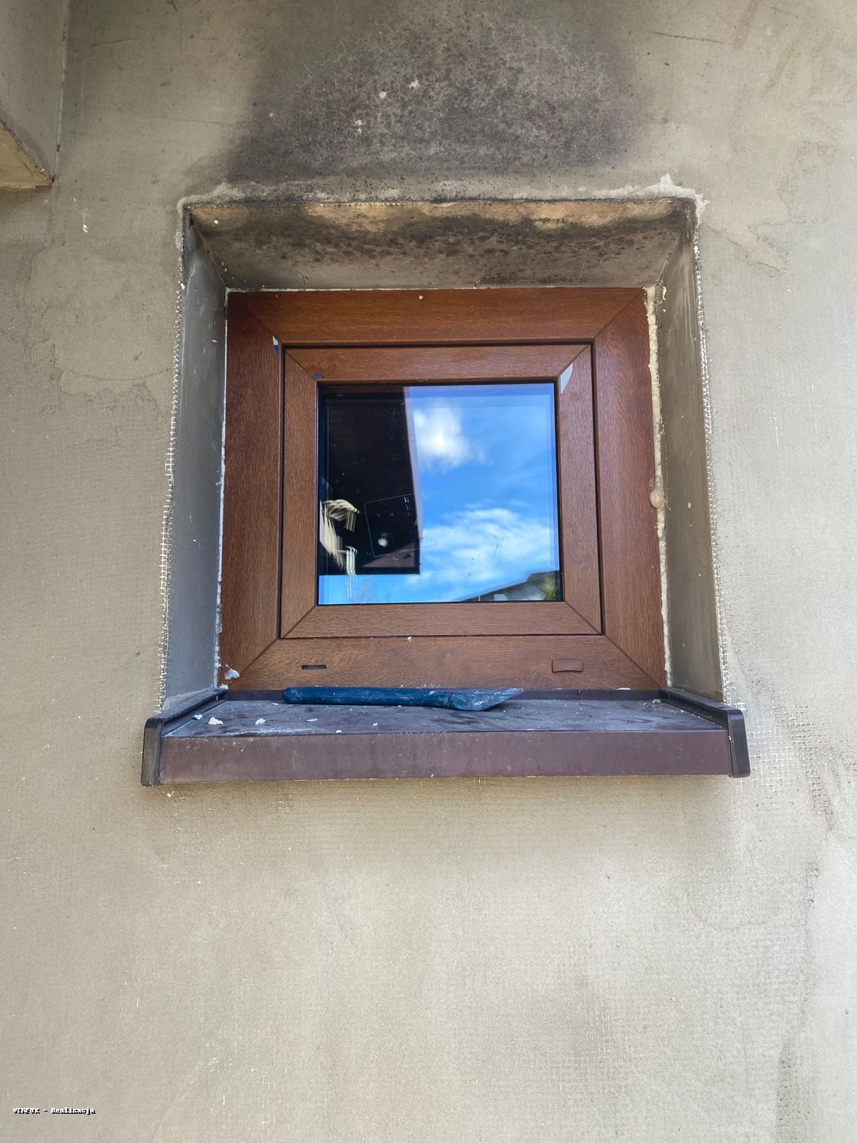 Montaż okna z brązową ramą w surowej elewacji. Widoczna szpachelka na parapecie i siatka elewacyjna. Okno odbija błękitne niebo z chmurami.