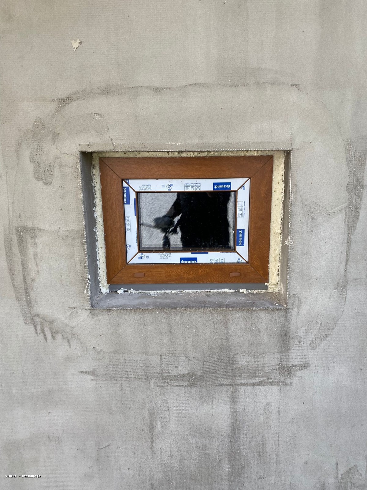 Brązowe okno z białymi ramami Deceuninck osadzone w surowej ścianie z widoczną pianką montażową i śladami po tynku. Widoczne odbicie osoby w szybie.