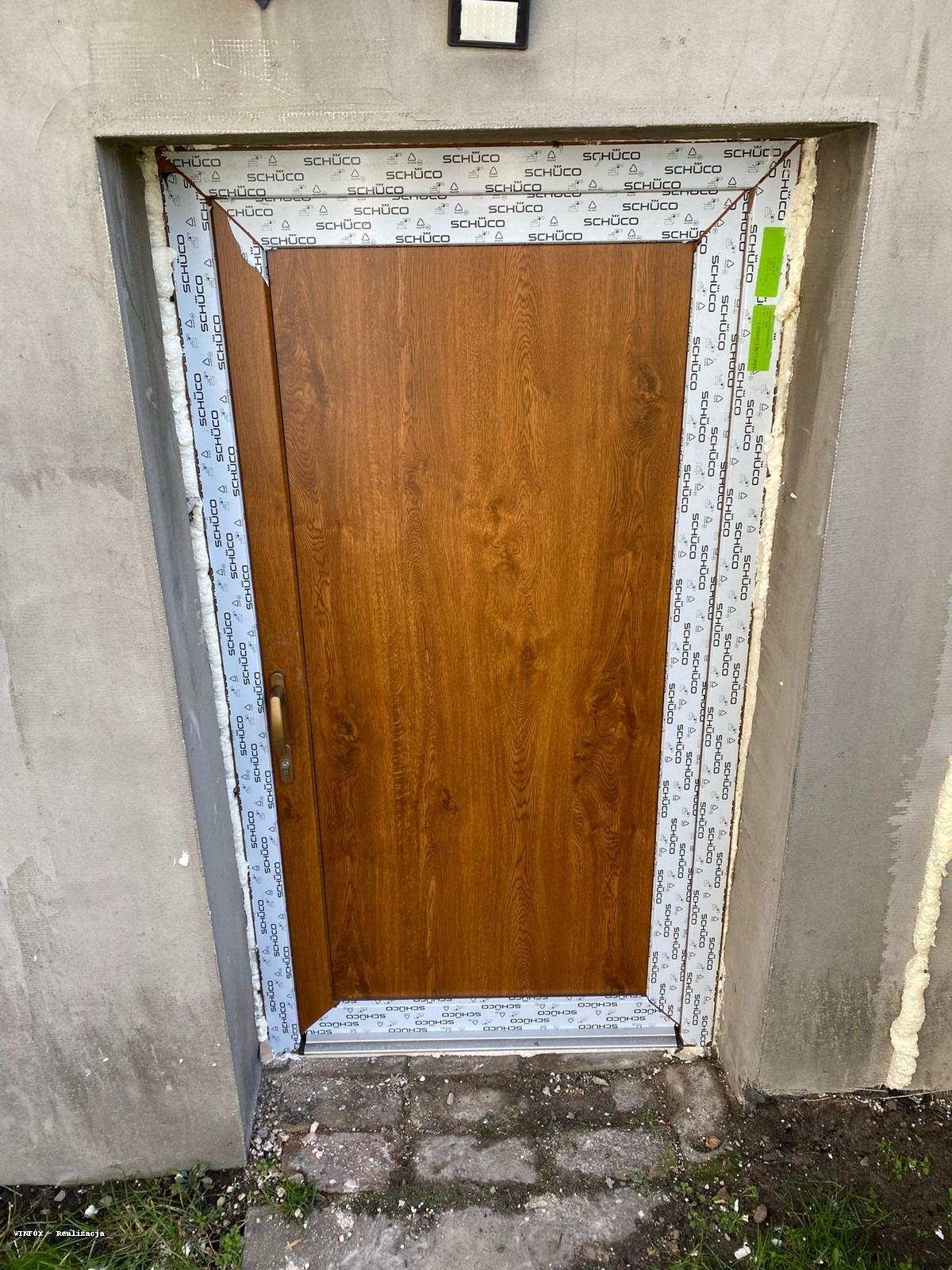 Brązowe drzwi zewnętrzne Schüco, zamontowane w surowym otworze, z widoczną pianką montażową i taśmą uszczelniającą. Widoczne logo producenta na ramie.