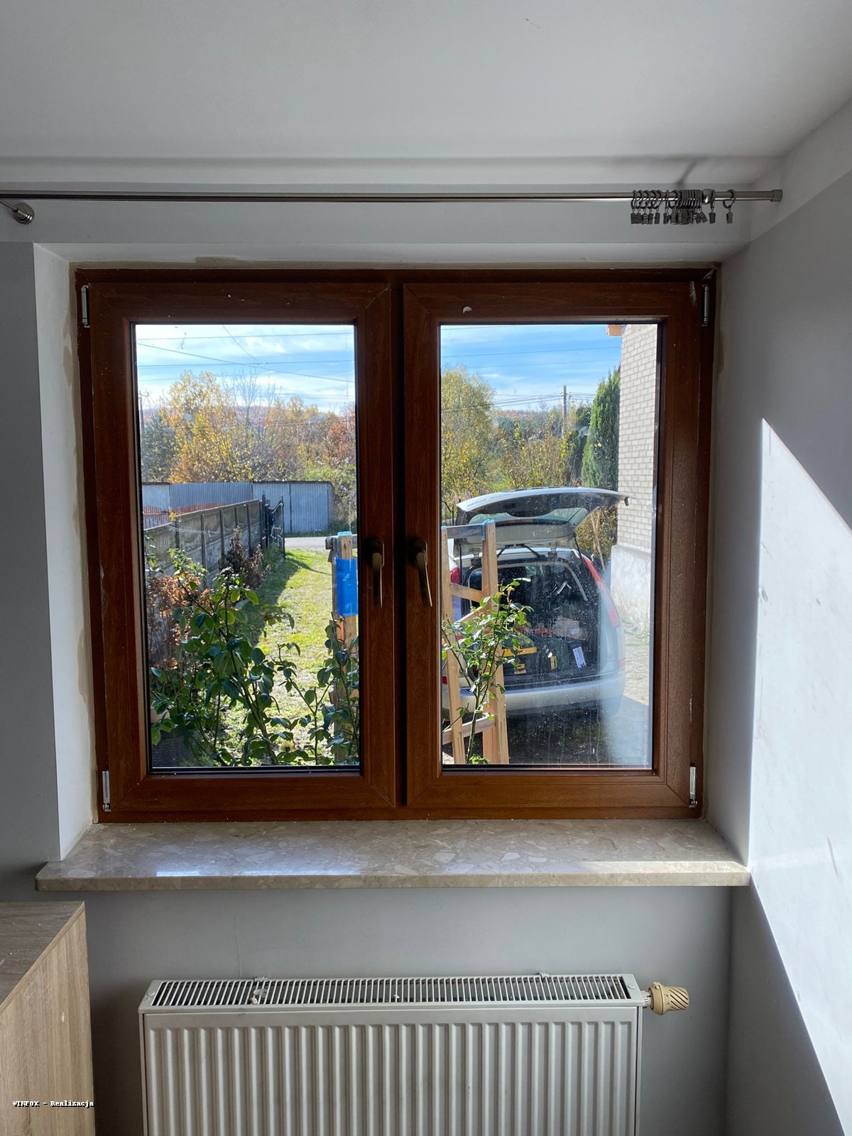 Dwuskrzydłowe okno z brązową ramą, widok na ogród i samochód z otwartym bagażnikiem. Wnętrze z grzejnikiem pod parapetem. Widoczne ślady po montażu.