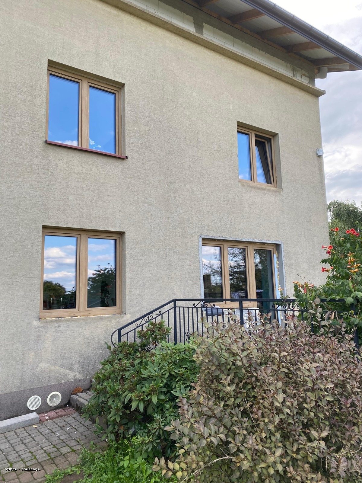 Elewacja budynku z beżowym tynkiem i oknami w drewnianych ramach. Widoczne schody z czarną balustradą i roślinność. Okno na górze jest uchylone.