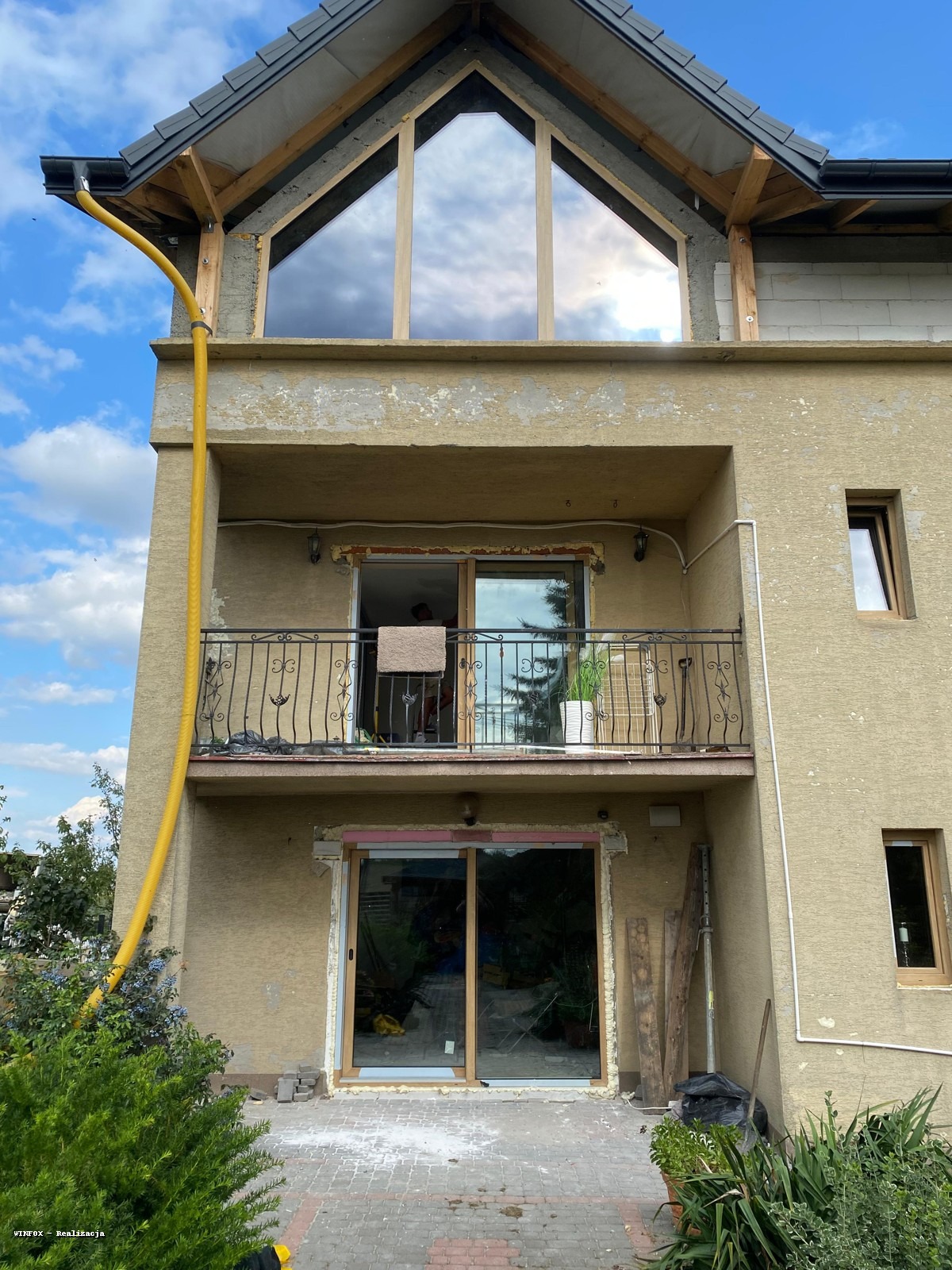 Dom w trakcie montażu stolarki okiennej i drzwiowej. Widoczne okno trójkątne na poddaszu, balkon z balustradą i częściowo zamontowane drzwi przesuwne na parterze.