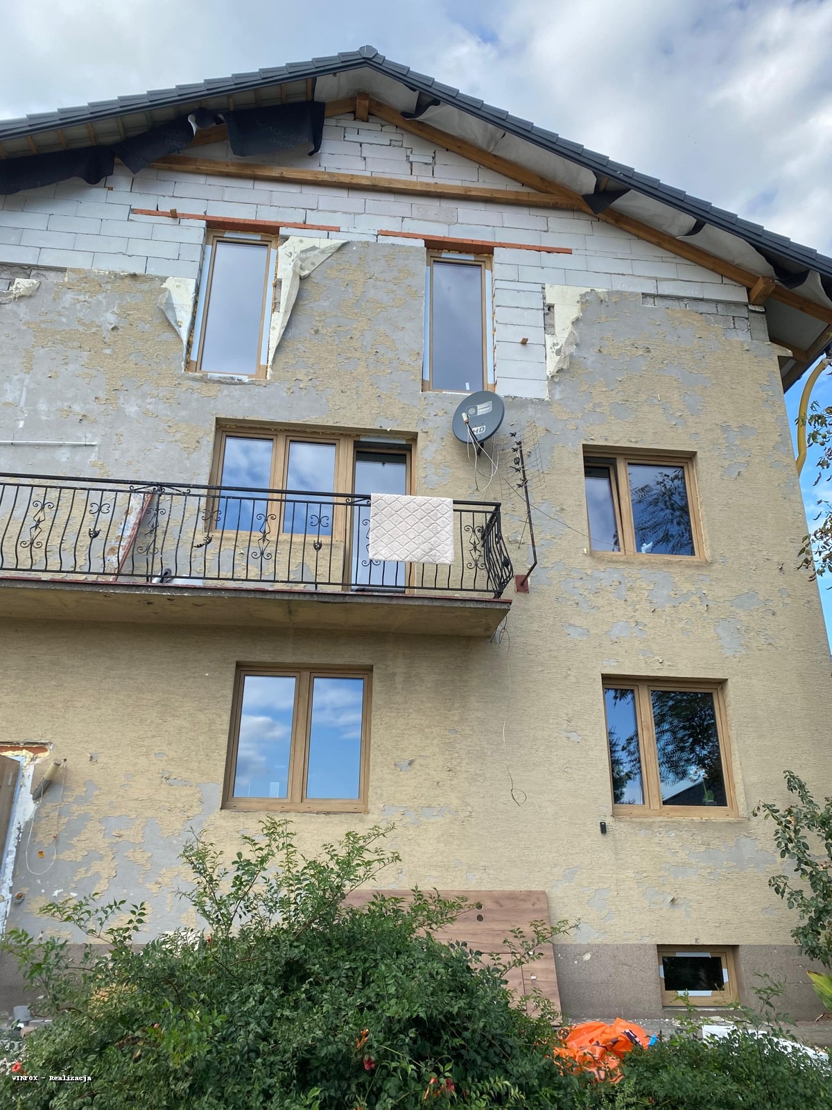 Dom w trakcie wymiany okien. Widoczne nowe okna z brązowymi ramami, fragmenty ocieplenia i elewacji w trakcie prac. Na balkonie suszy się ręcznik.