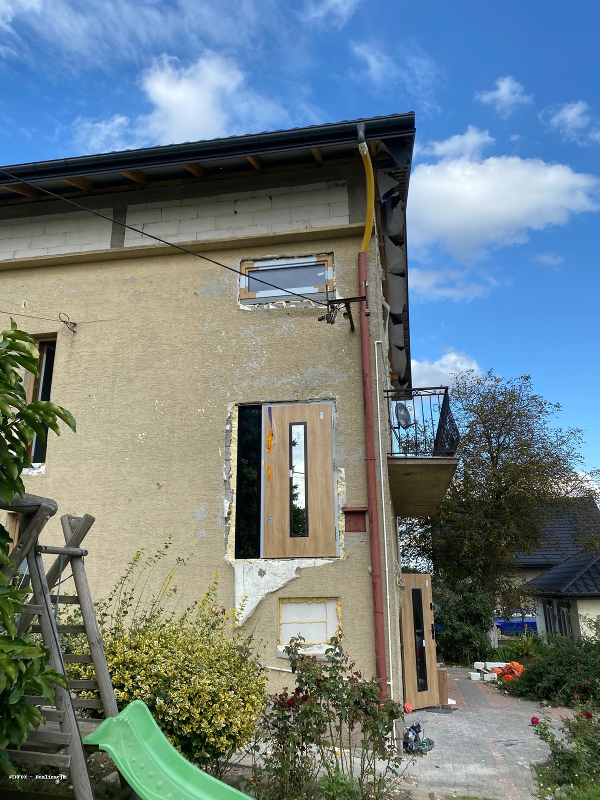 Montaż drzwi zewnętrznych w domu jednorodzinnym. Widoczne przygotowane otwory okienne i drzwiowe. Prace w toku, elewacja częściowo uszkodzona. Słoneczny dzień.