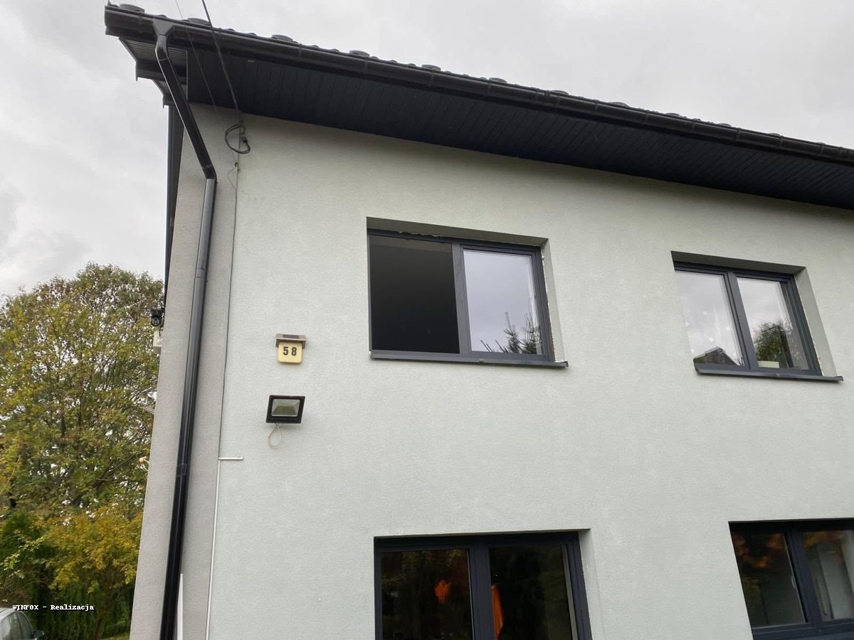 Elewacja domu z zamontowanymi oknami PCV w grafitowych ramach. Widoczne okna na parterze i piętrze, numer domu 58 i oświetlenie zewnętrzne.
