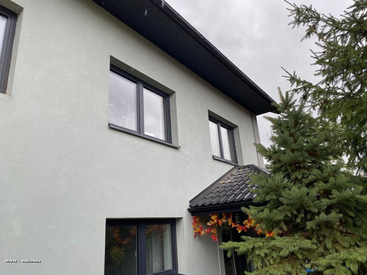 Elewacja domu z szarymi oknami, widok z dołu. Dekoracja jesienna nad wejściem, iglak po prawej. Szara elewacja i ciemna podbitka dachowa.