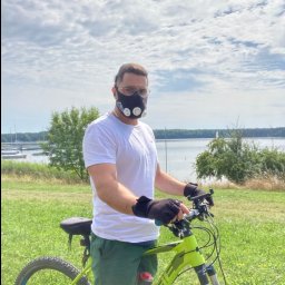 Marcel Alsahwi- Trener Personalny - Mężczyzna w masce treningowej na rowerze górskim, na tle jeziora i zieleni. Trening na świeżym powietrzu z użyciem specjalistycznego sprzętu.