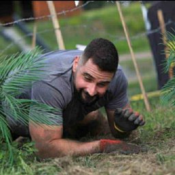 Marcel Alsahwi- Trener Personalny - Uśmiechnięty mężczyzna czołga się pod drutem kolczastym podczas ekstremalnego biegu z przeszkodami, ubrany w rękawice, z roślinnością w tle.