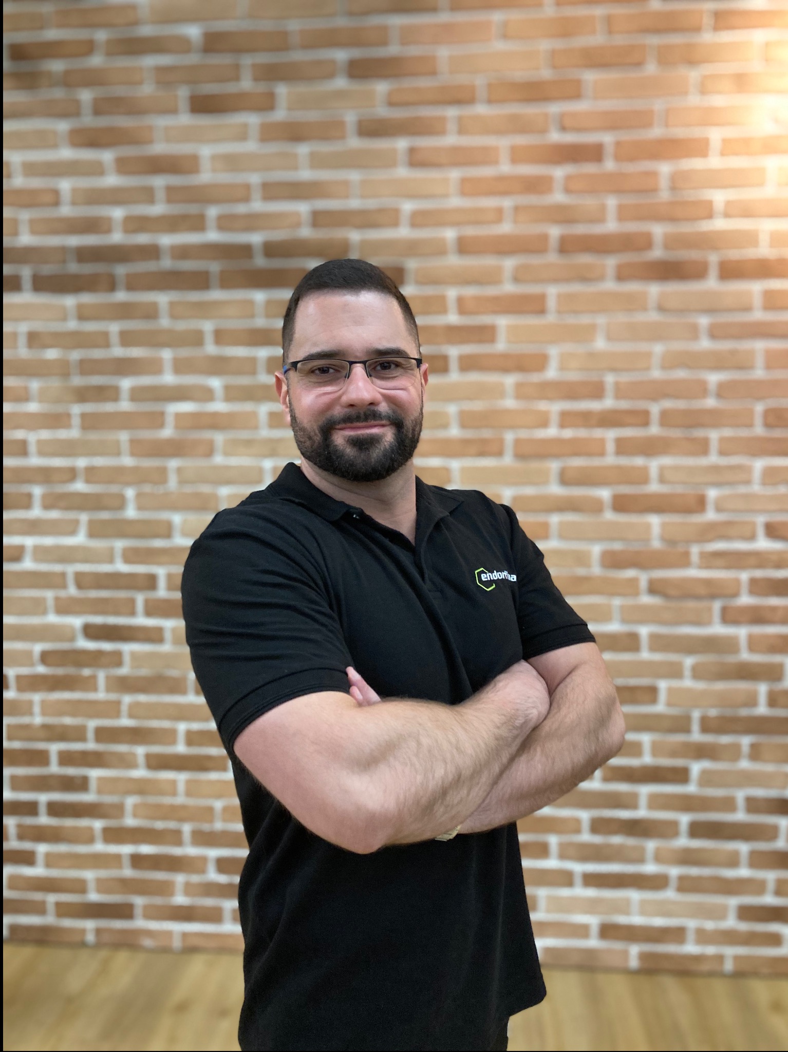Uśmiechnięty mężczyzna w okularach, z brodą i w czarnej koszulce polo, stoi na tle ceglanej ściany z założonymi rękami. Widoczny logotyp na koszulce.