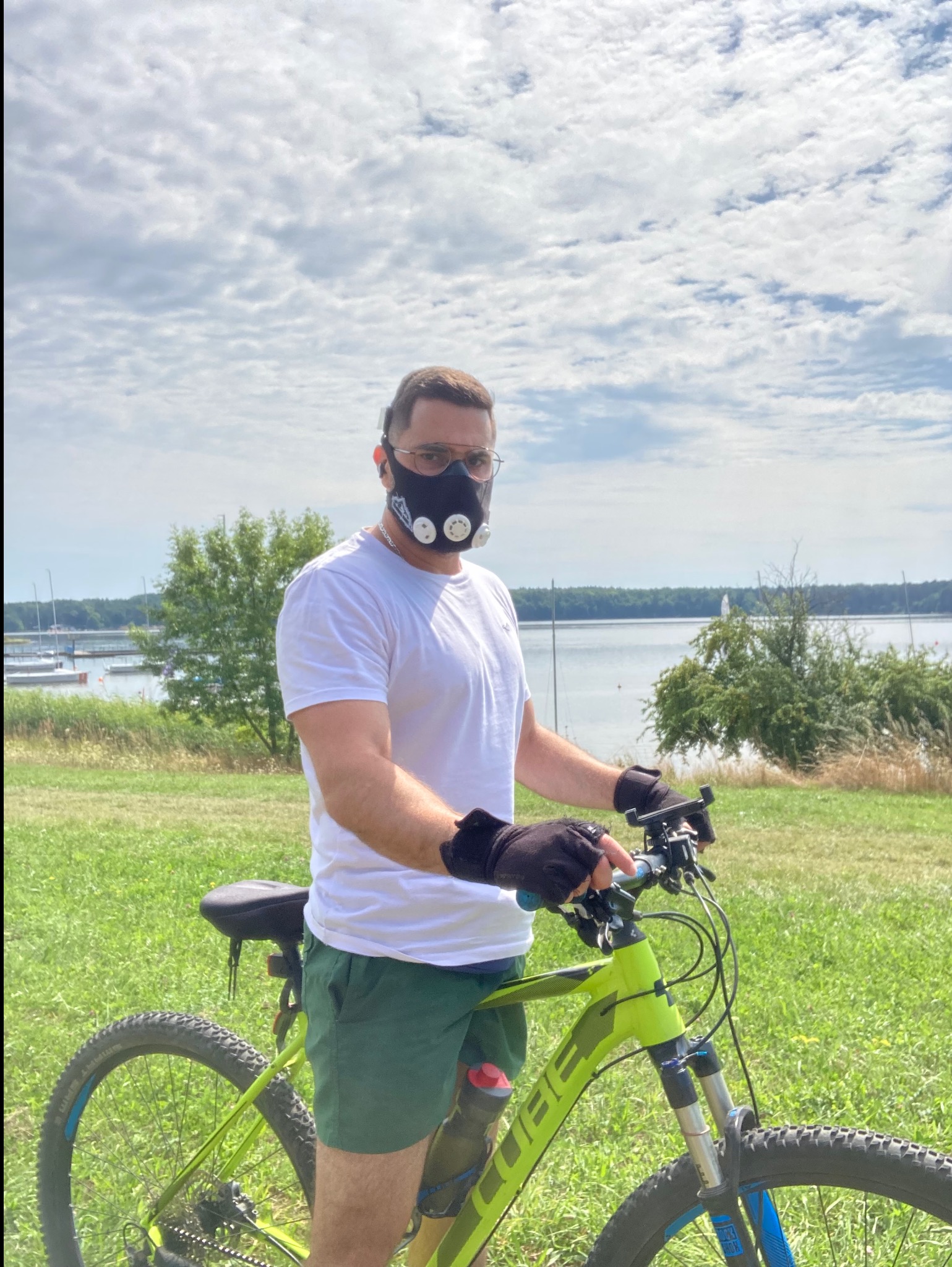 Mężczyzna w masce treningowej na rowerze górskim, na tle jeziora i zieleni. Trening na świeżym powietrzu z użyciem specjalistycznego sprzętu.