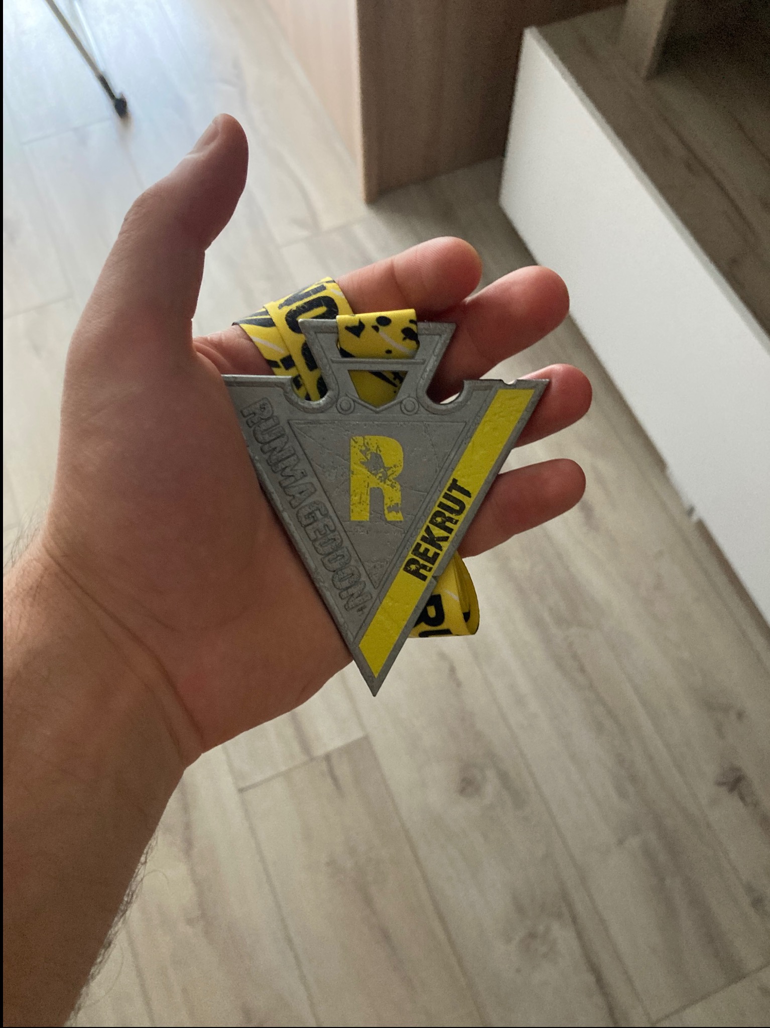 Dłoń trzyma medal Runmageddon Rekrut z żółtą wstążką. Medal w kształcie strzałki, szaro-żółty, z napisem 'REKRUT' i 'RUNMAGEDDON'.