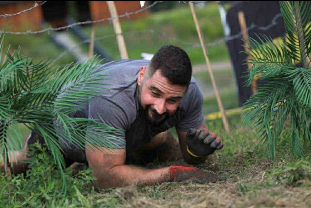 Uśmiechnięty mężczyzna czołga się pod drutem kolczastym podczas ekstremalnego biegu z przeszkodami, ubrany w rękawice, z roślinnością w tle.