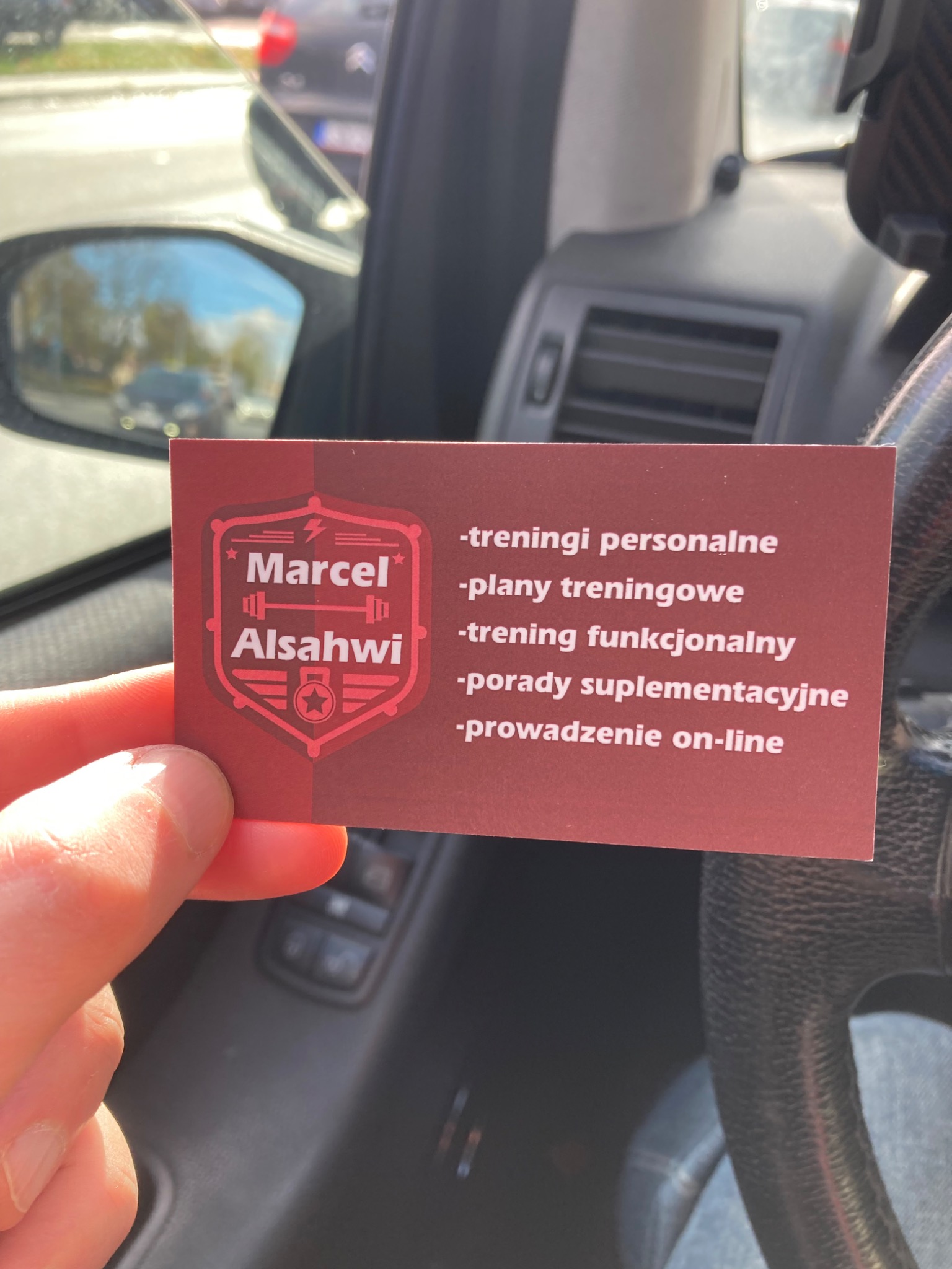 Wnętrze auta: dłoń trzyma wizytówkę Marcela Alsahwi z ofertą treningów personalnych, planów treningowych, treningu funkcjonalnego, porad suplementacyjnych i prowadzenia on-line.