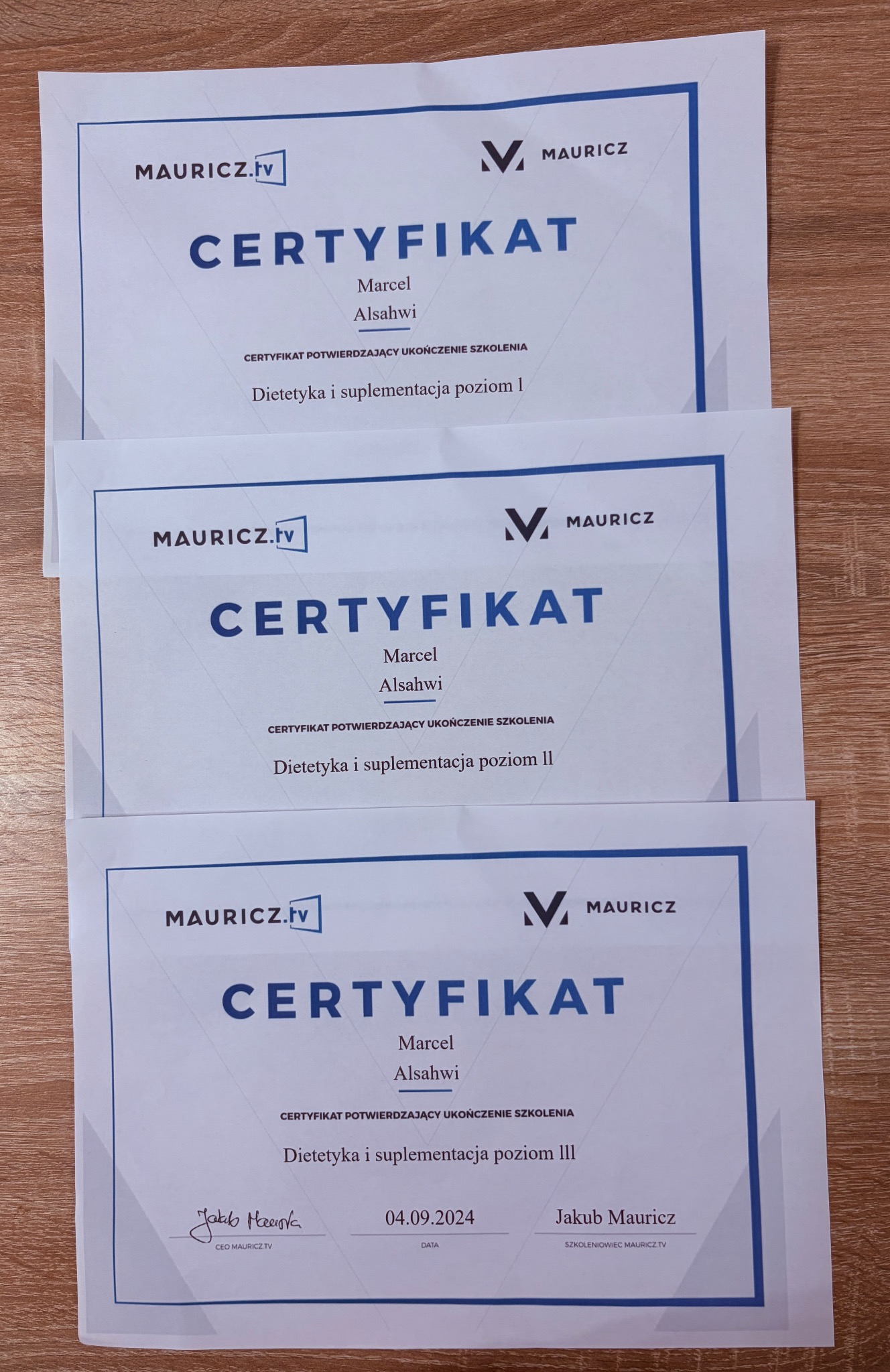Trzy certyfikaty ukończenia szkolenia z dietetyki i suplementacji (poziom I, II, III) Marcela Alsahwi, sygnowane przez Mauricz.tv, leżące na drewnianym podłożu.