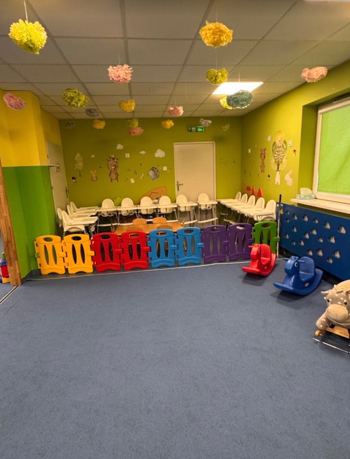 Kolorowa sala zabaw z krzesełkami do karmienia, niskim płotkiem i zabawkami na biegunach. Dekoracje sufitowe w kształcie pomponów. Ściany zdobią naklejki ze zwierzętami.