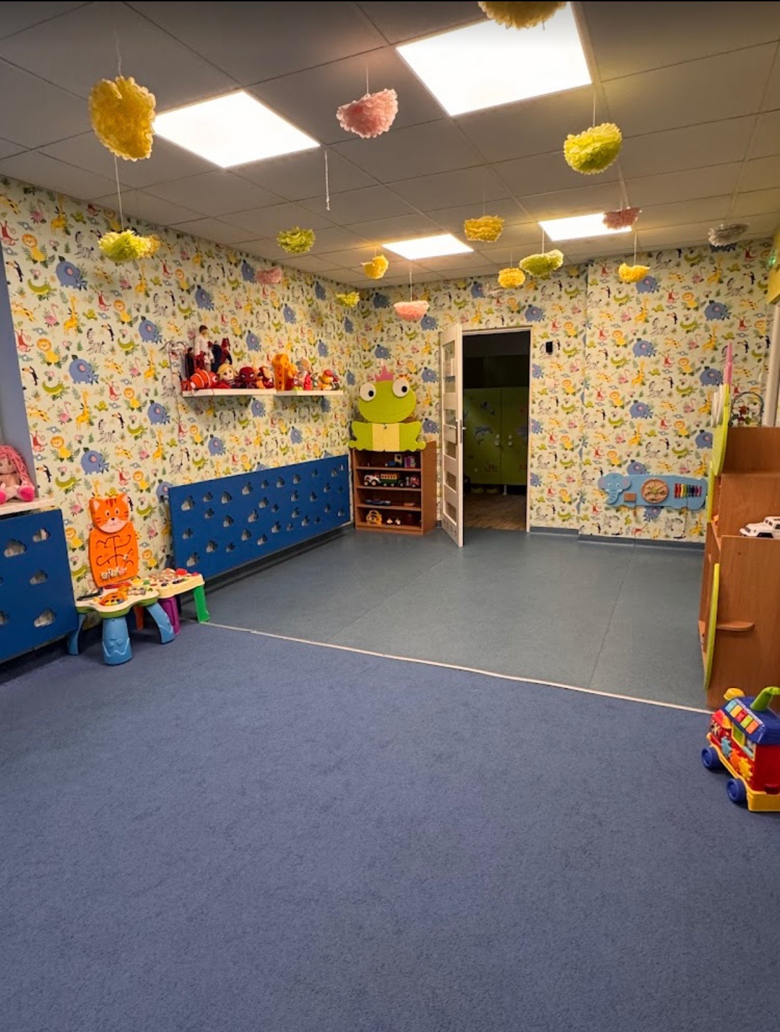 Kolorowa sala zabaw z motywem zwierzęcym na ścianach, wyposażona w niskie meble, zabawki i dekoracje podwieszone pod sufitem. Widoczny fragment podłogi z niebieską wykładziną.