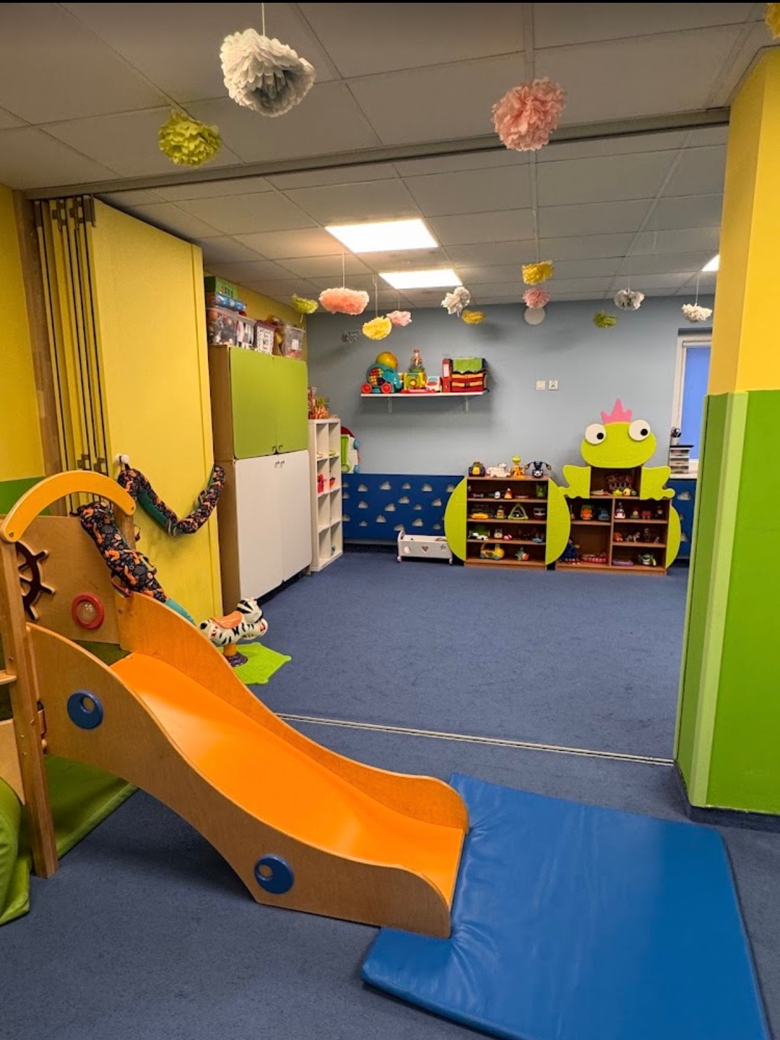 Kolorowa sala zabaw z pomarańczową zjeżdżalnią, regałami w kształcie żaby i wiszącymi ozdobami z bibuły. Niebieska wykładzina i pastelowe ściany tworzą przyjazną atmosferę.