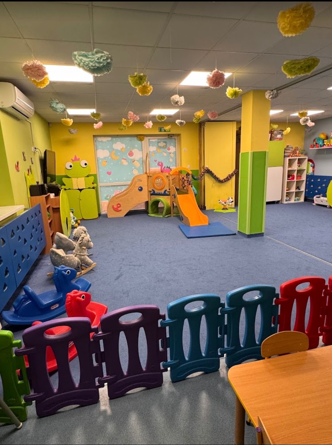 Kolorowa sala zabaw z niskim ogrodzeniem, zjeżdżalniami i dekoracjami pod sufitem. Widoczne zabawki i meble dostosowane do dziecięcego wzrostu. Przytulne, bezpieczne otoczenie.