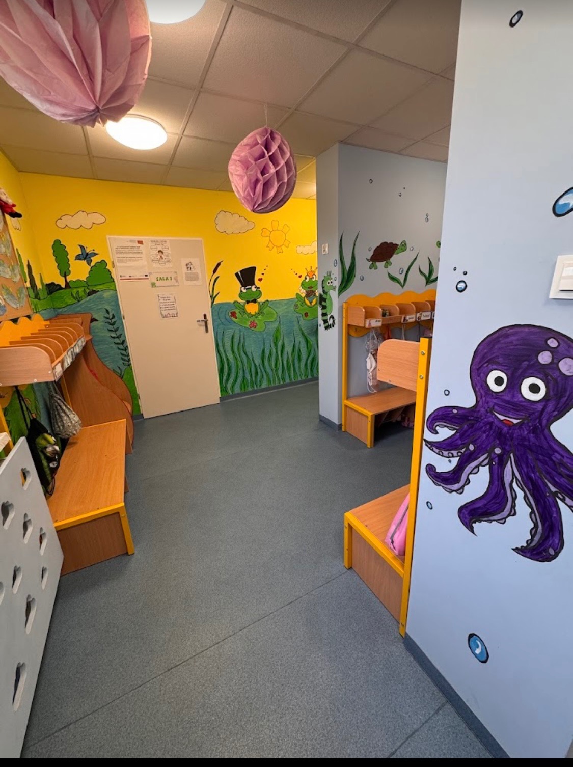 Kolorowy korytarz w placówce edukacyjnej z rysunkami zwierząt na ścianach, szafkami i ławkami dla dzieci oraz dekoracjami wiszącymi pod sufitem.