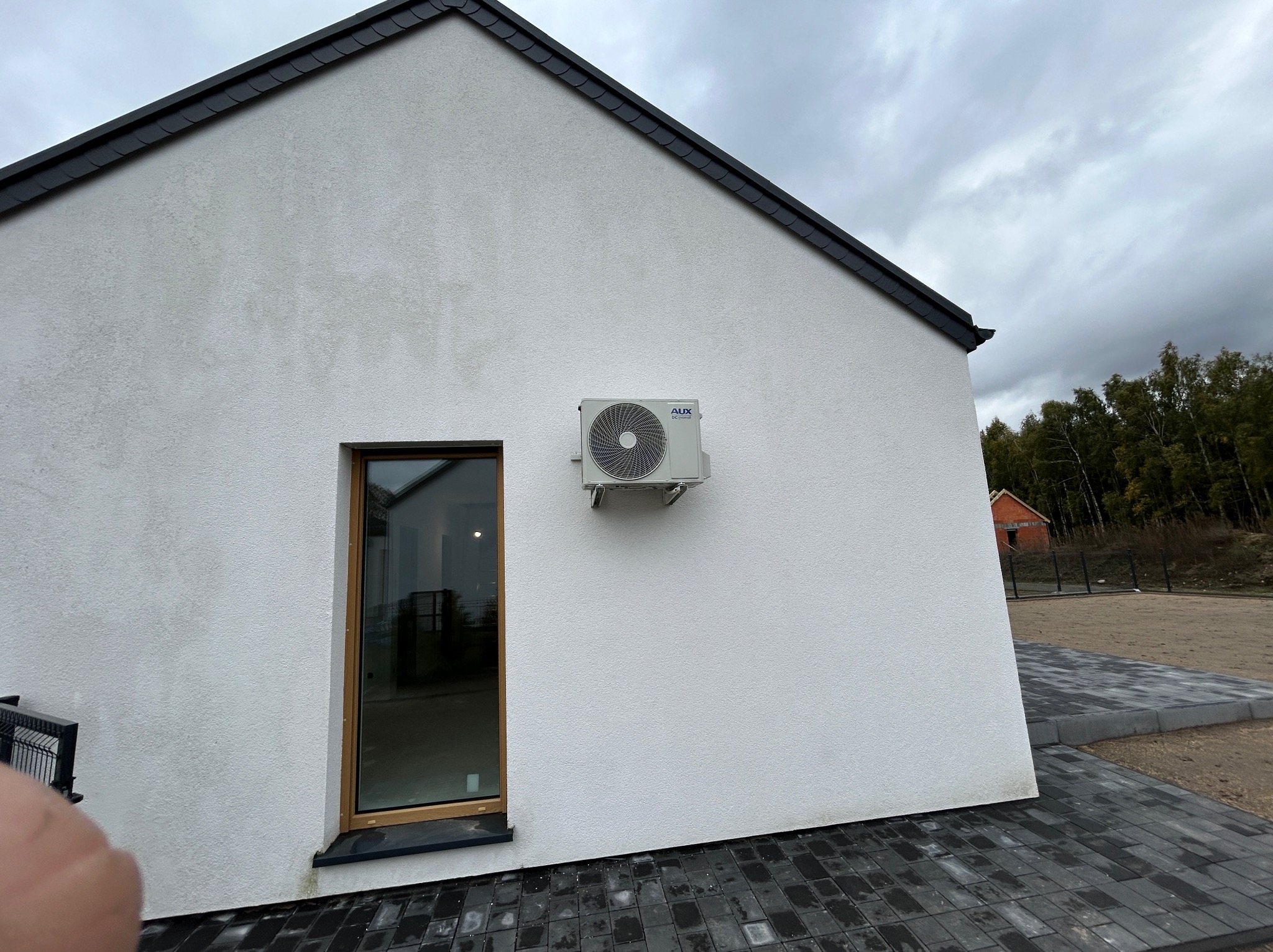Biała ściana domu z zamontowaną jednostką zewnętrzną klimatyzacji AUX, widok z boku, okno z drewnianą ramą, kostka brukowa, pochmurne niebo.