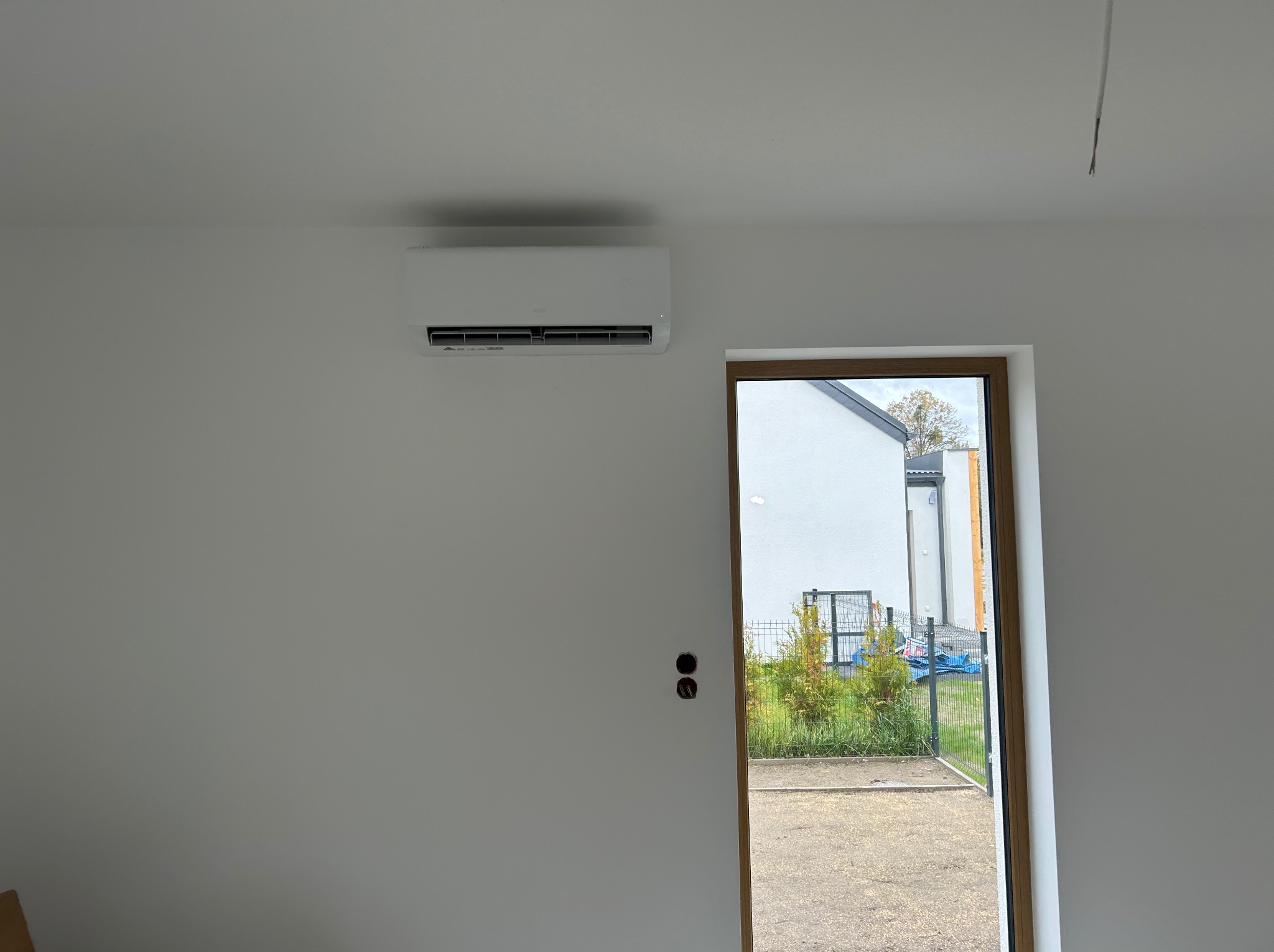 Biała klimatyzacja na ścianie w jasnym wnętrzu, widok przez otwarte drzwi na zewnątrz. Minimalistyczny design, świeżo pomalowane ściany, widoczne otwory na kable.