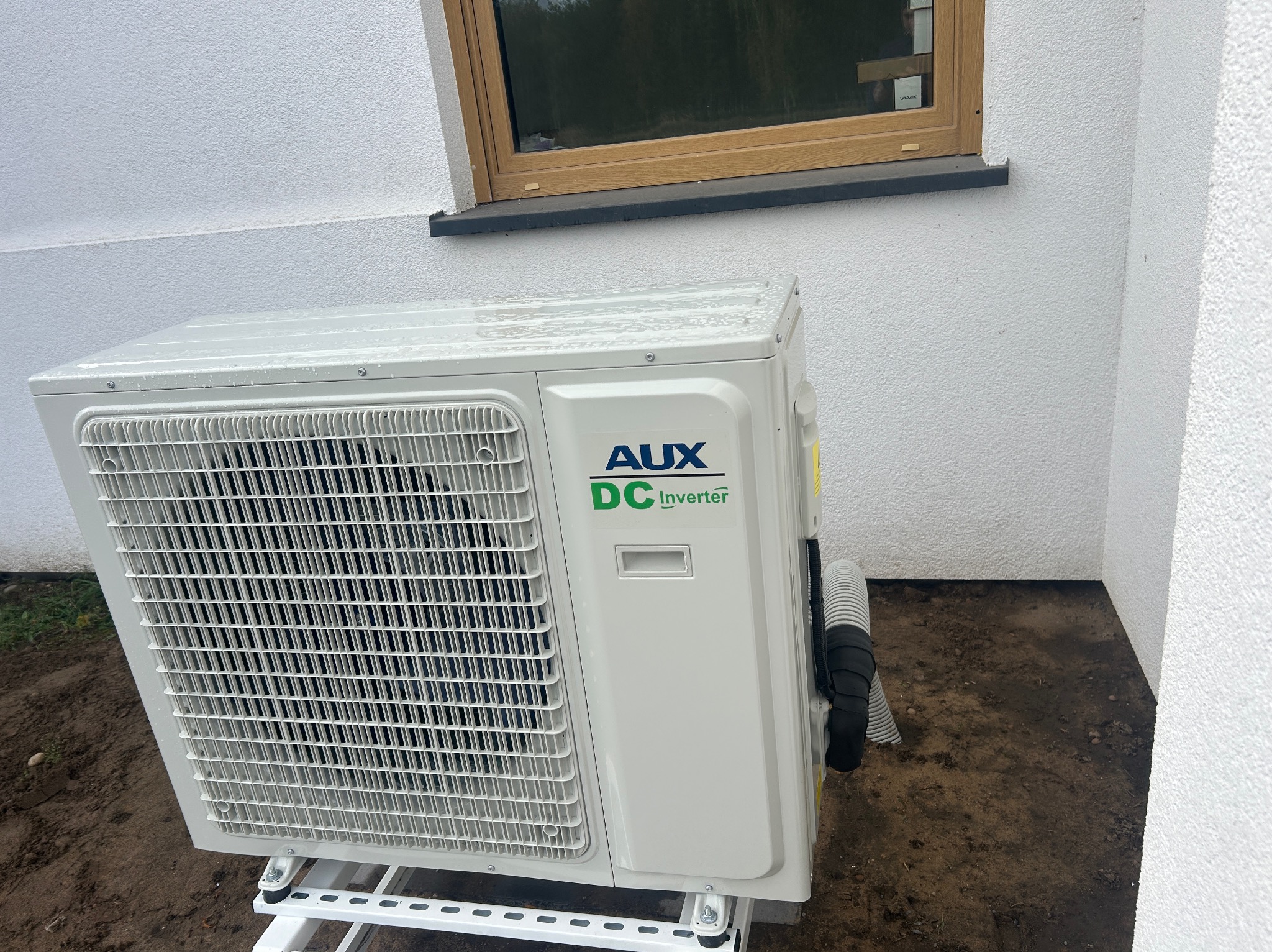 Biała jednostka zewnętrzna klimatyzacji AUX DC Inverter zamontowana na białym stelażu przy ścianie budynku z oknem. Widoczne przewody i ziemiste podłoże.