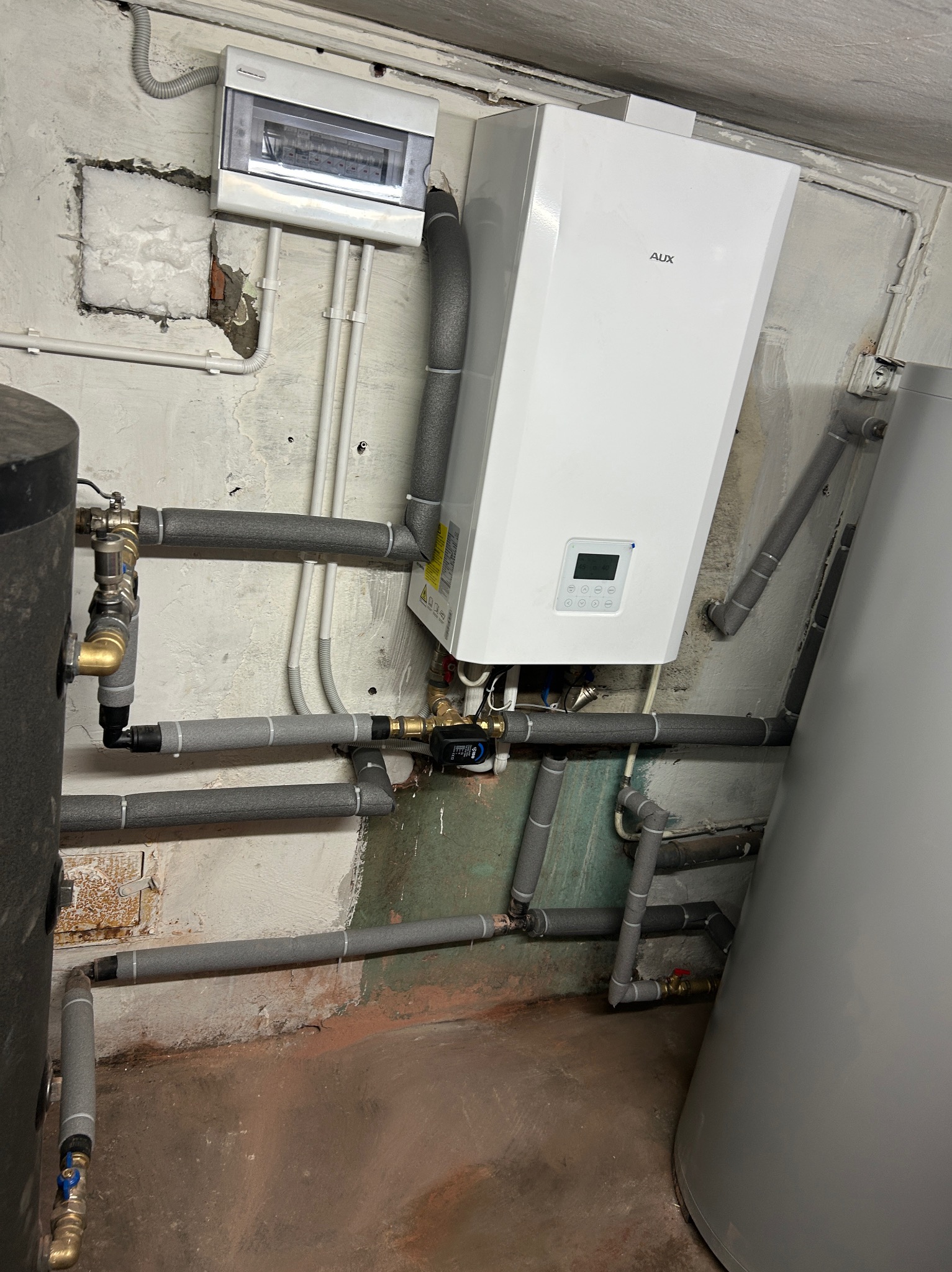 Biały piec grzewczy AUX zamontowany na ścianie z szarymi rurami w izolacji, widoczny zbiornik i skrzynka elektryczna. Instalacja w piwnicy.