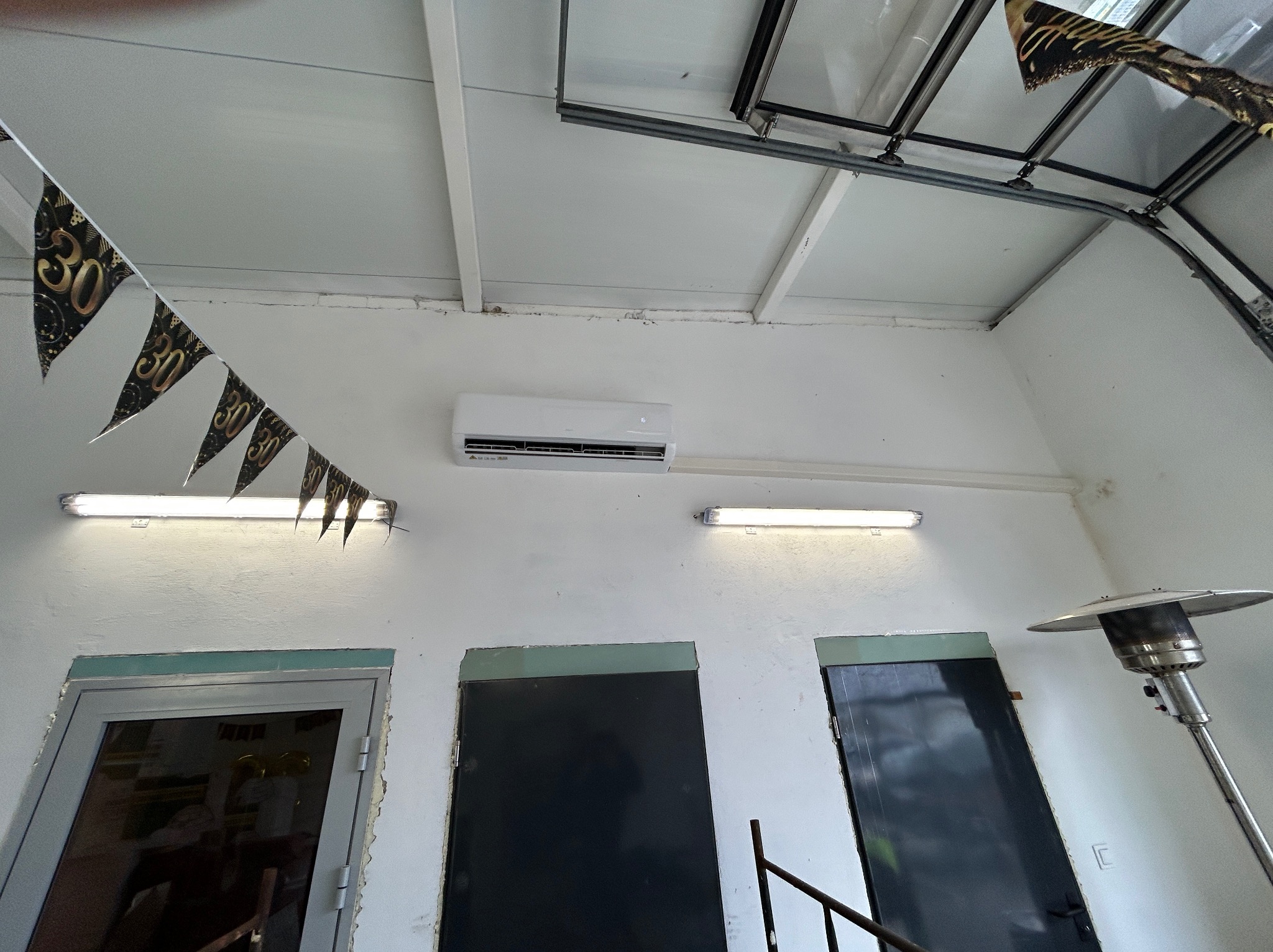 Biała klimatyzacja ścienna w pomieszczeniu z trzema drzwiami i dekoracją urodzinową, oświetlona lampami. Widok z dołu.