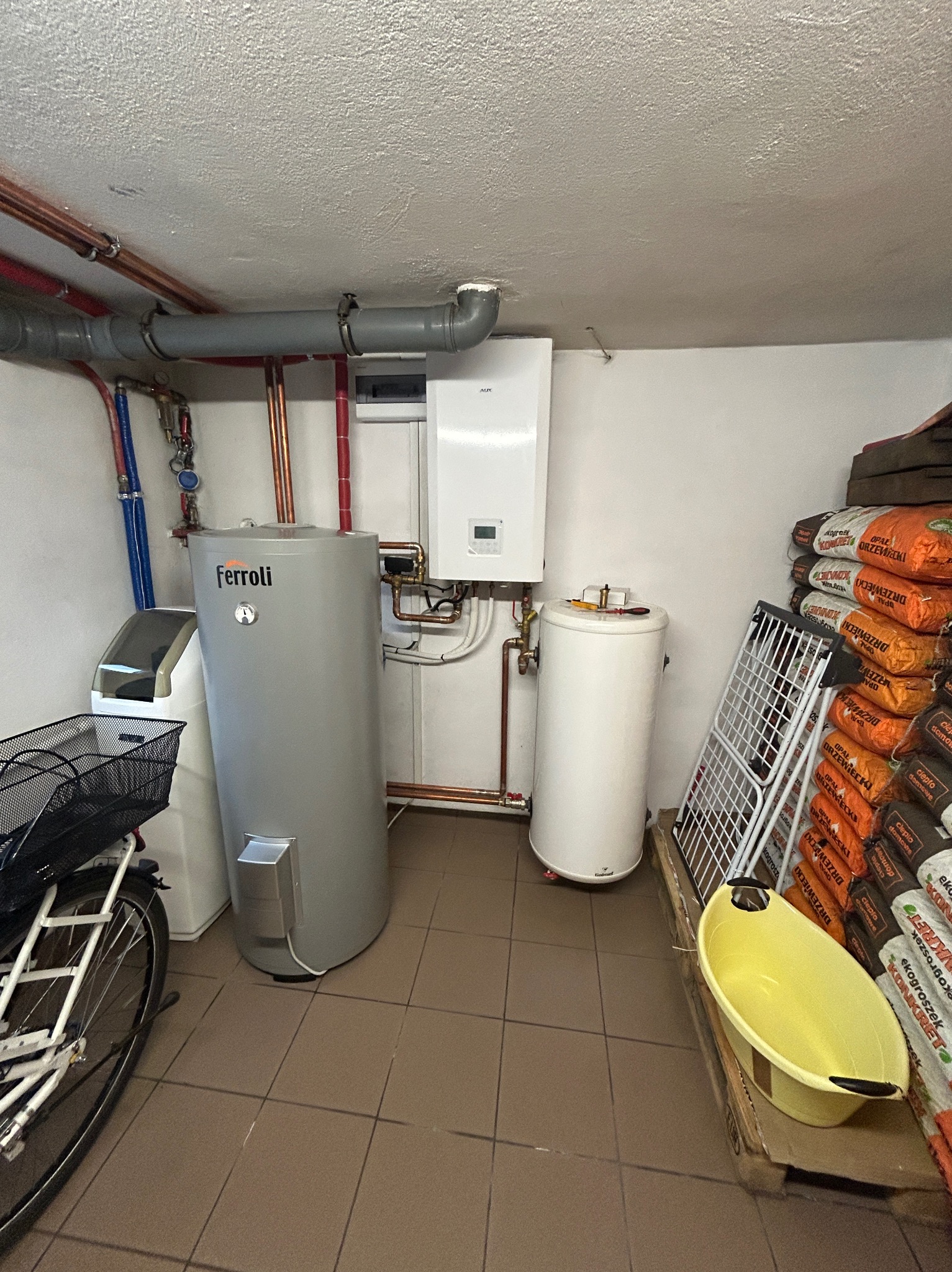 Kotłownia z szarym zasobnikiem Ferroli, białym bojlerem i workami opału. Widoczne rury instalacyjne i rower. Ujęcie w pomieszczeniu z brązowymi płytkami.