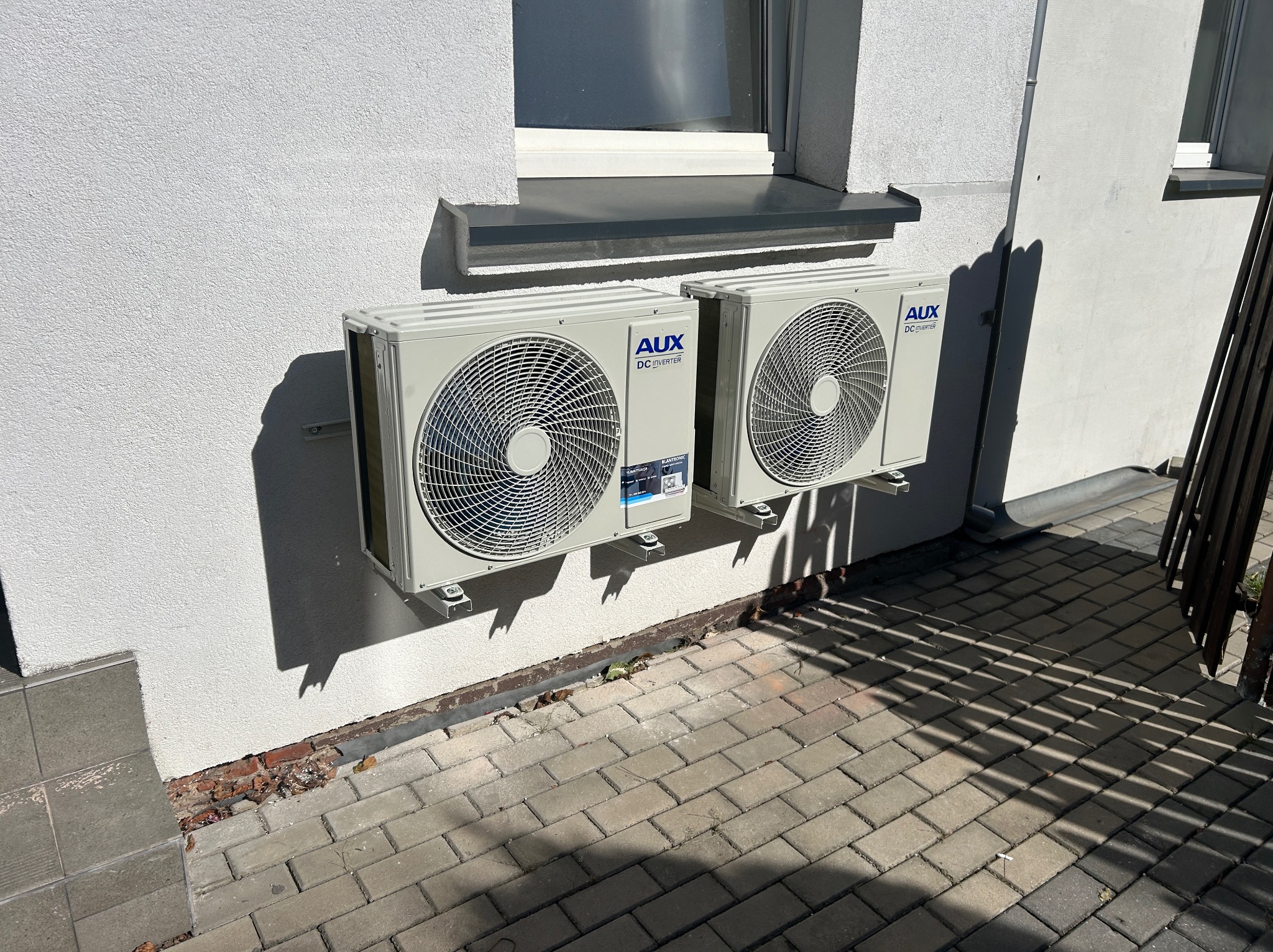Dwa klimatyzatory AUX DC Inverter zamontowane na białej elewacji budynku, widoczne z poziomu chodnika z kostki brukowej w słoneczny dzień.