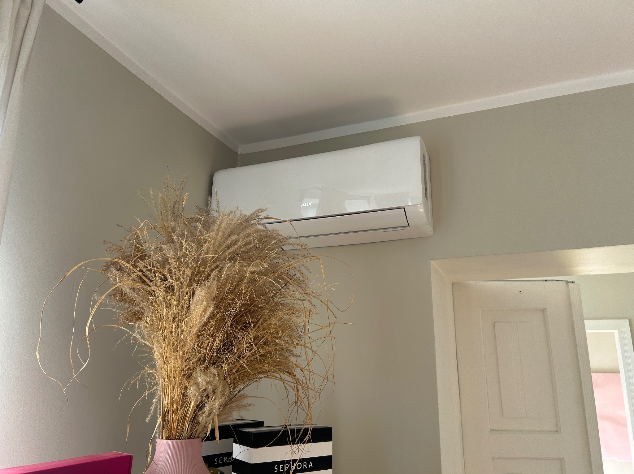 Biały klimatyzator AUX na ścianie w pokoju, obok drzwi i dekoracji z suszonych traw w wazonie. Widok z dołu, neutralne kolory ścian.