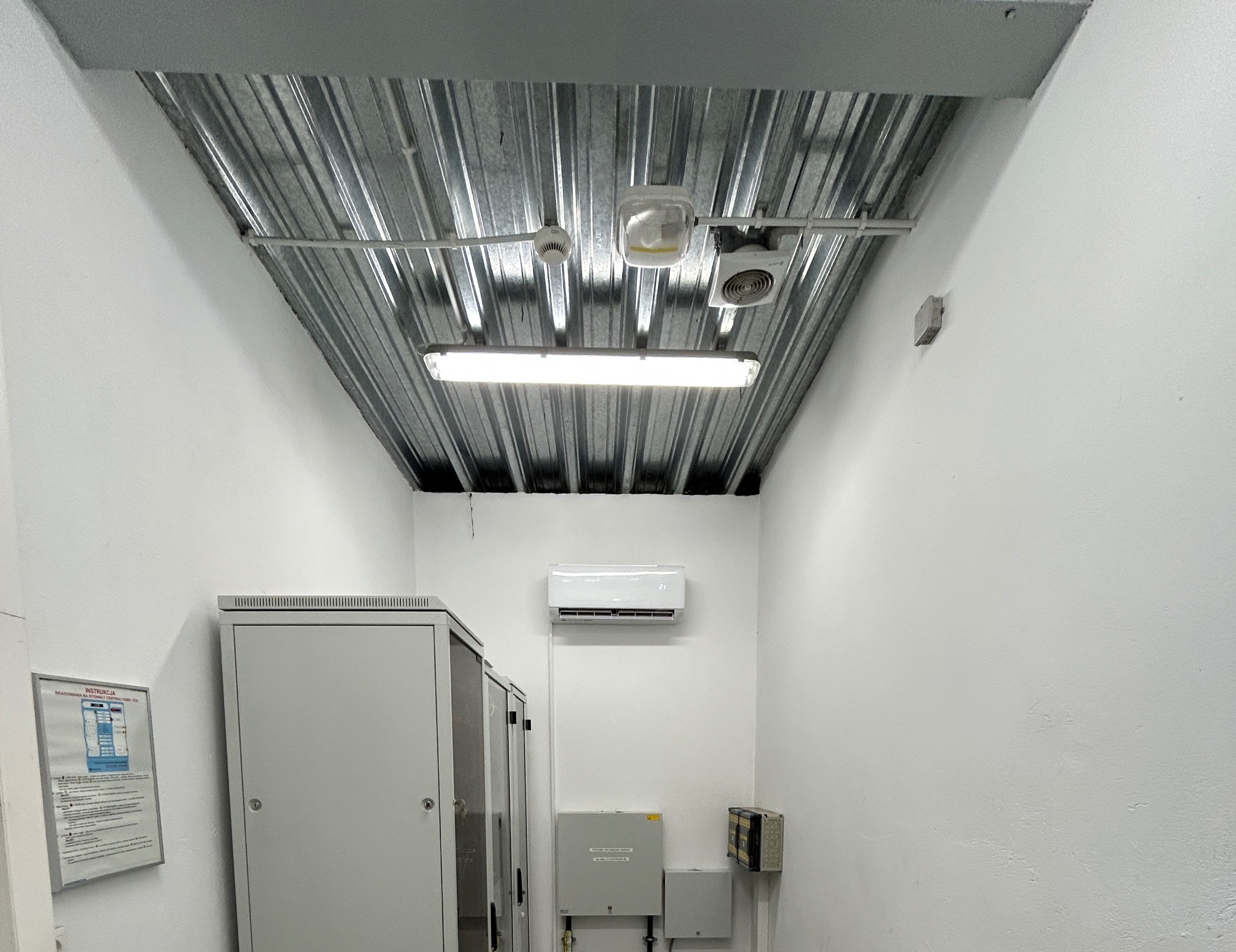 Białe pomieszczenie techniczne z zamontowaną klimatyzacją, metalowym sufitem, oświetleniem i szafami elektrycznymi. Widoczny czujnik dymu i wentylator.