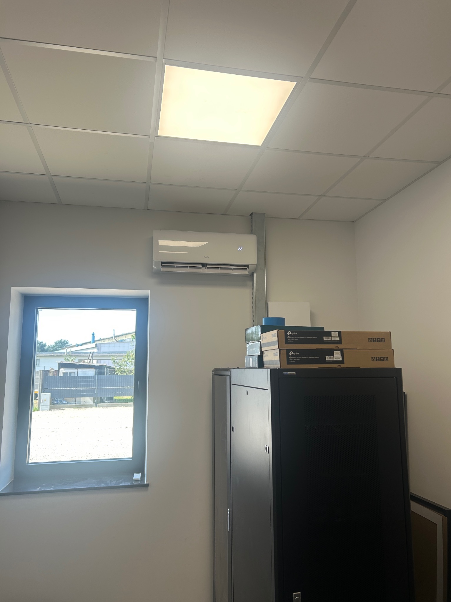 Biała klimatyzacja ścienna zamontowana w pomieszczeniu biurowym, obok okna i szafy rack z pudełkami. Widoczne oświetlenie sufitowe.