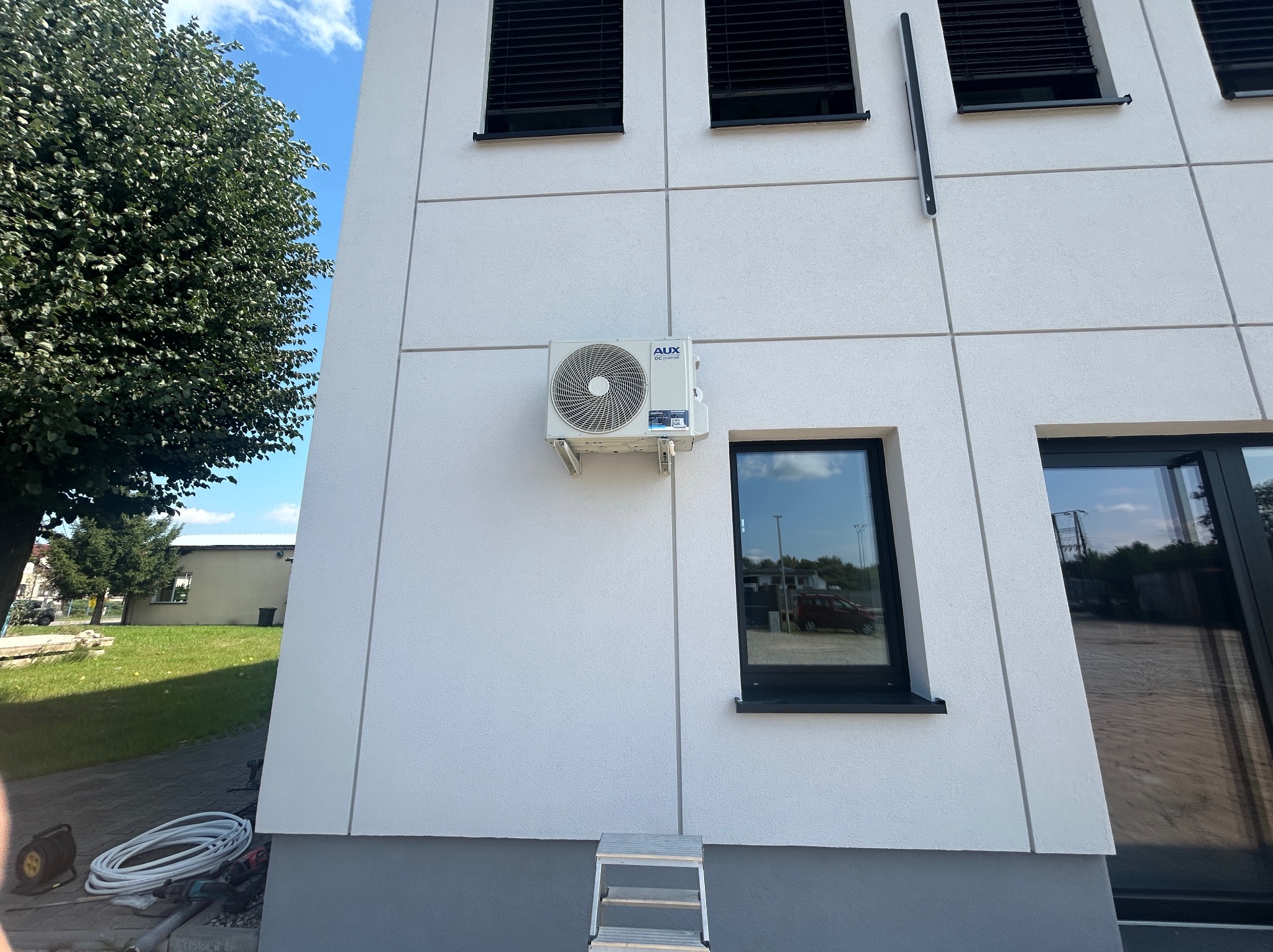 Biała jednostka klimatyzacji AUX zamontowana na elewacji budynku z szarą podmurówką. Obok okno z czarną ramą i odbiciem otoczenia. Widoczne narzędzia i drabina.